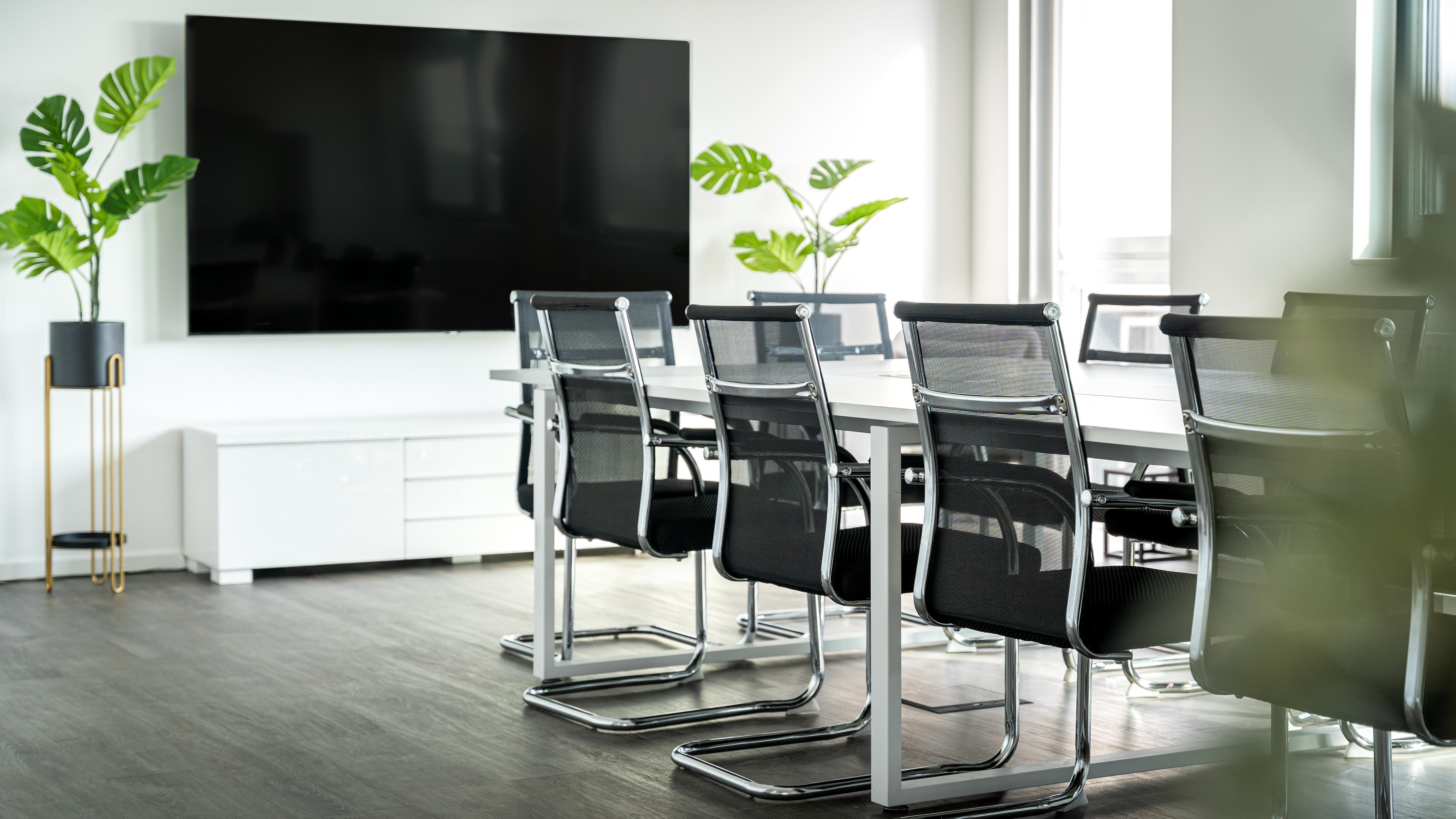 Ein moderner Konferenzraum mit einem großen Fernseher an der Wand, einem langen Tisch und sechs schwarzen Bürostühlen. Im Hintergrund stehen zwei große Pflanzen in Töpfen.
