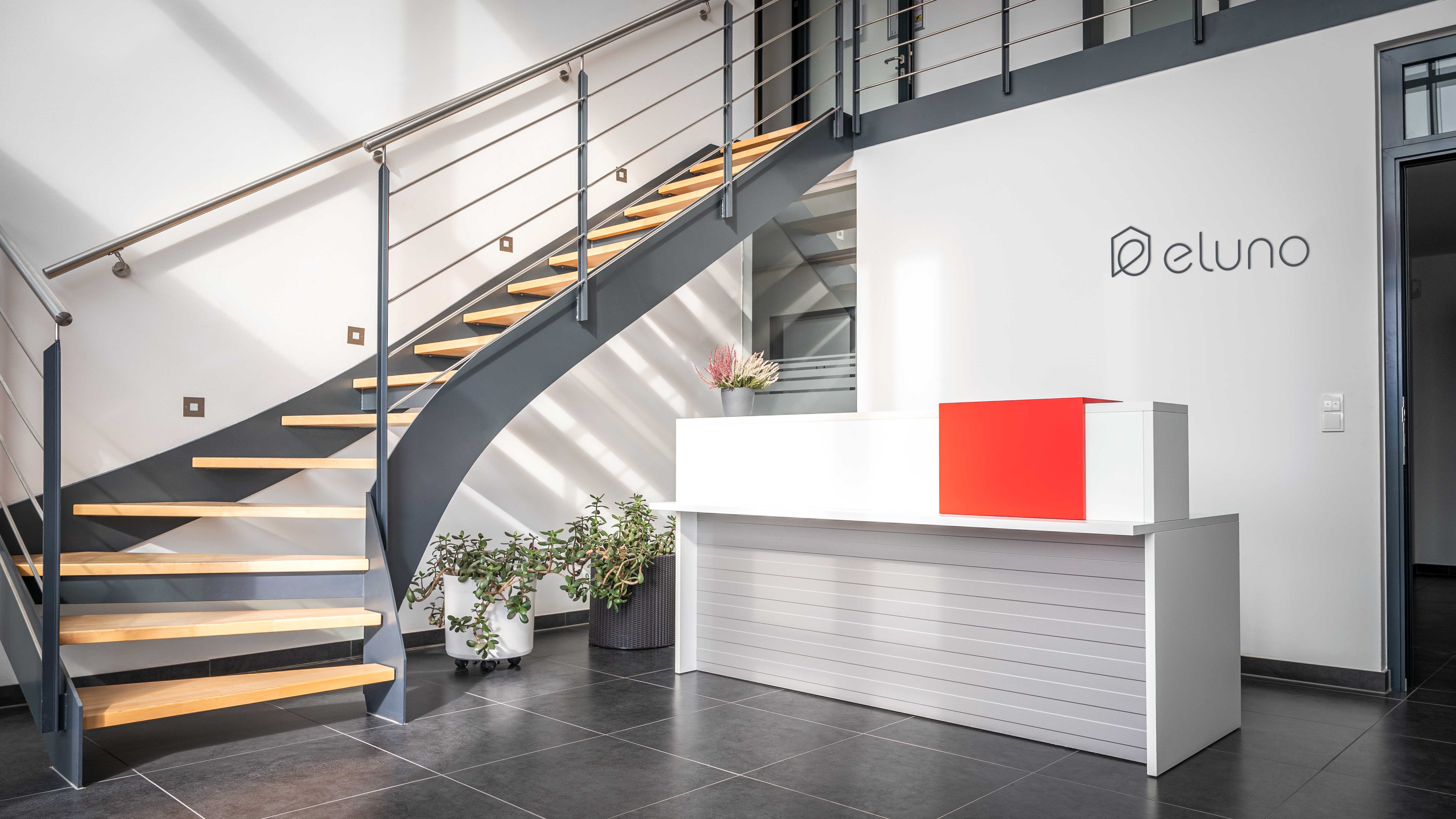 Moderne Empfangshalle mit einer geschwungenen Treppe aus Holz und Metall. Ein Empfangstresen in Weiß mit einem roten Element und Pflanzen im Hintergrund. An der Wand steht 'eluno'.