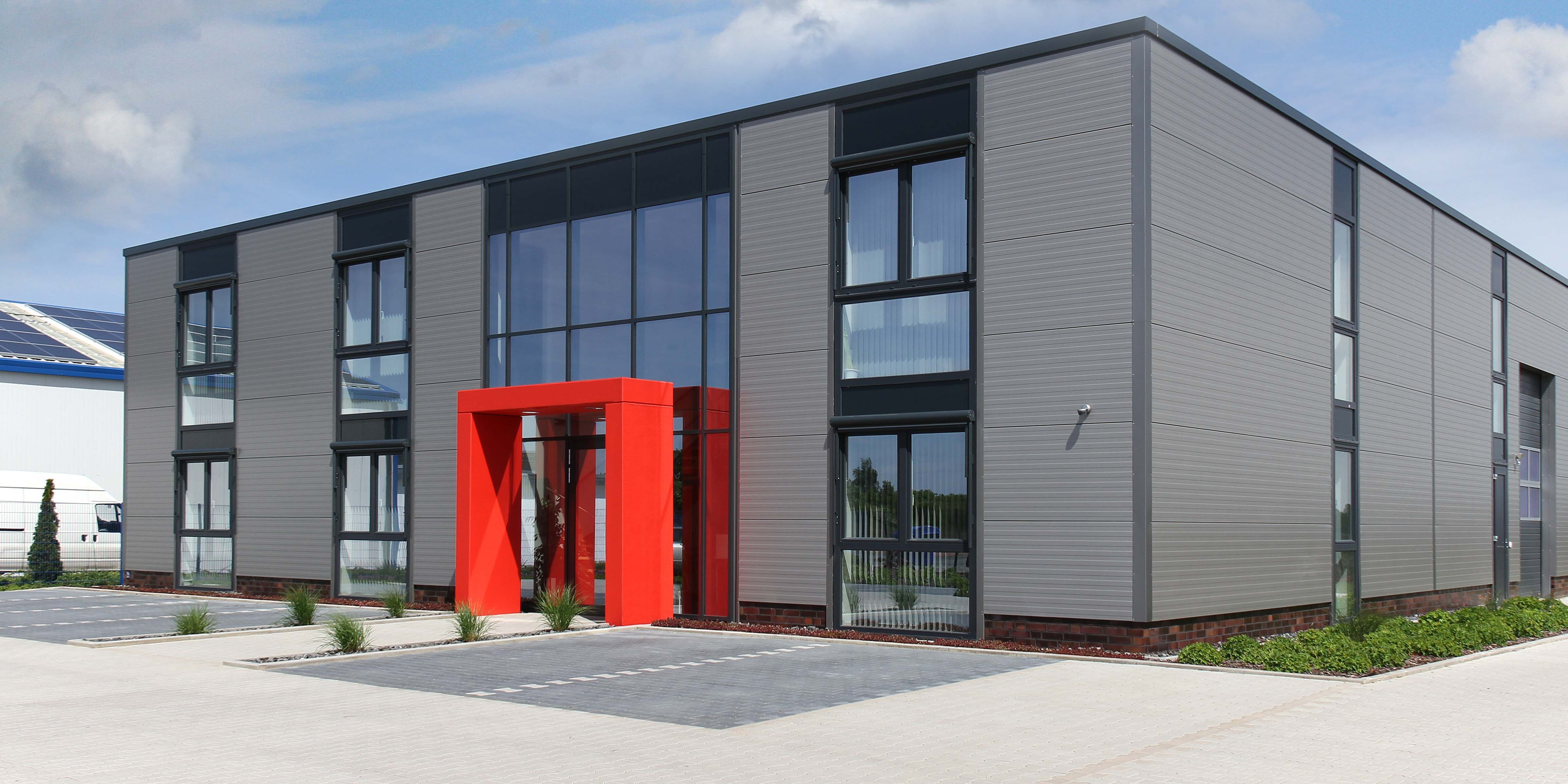 Moderne Gebäudeansicht mit grauer Fassade, großen Fenstern und einem roten Eingangsbereich. Umgeben von Rasen und Pflastersteinen.