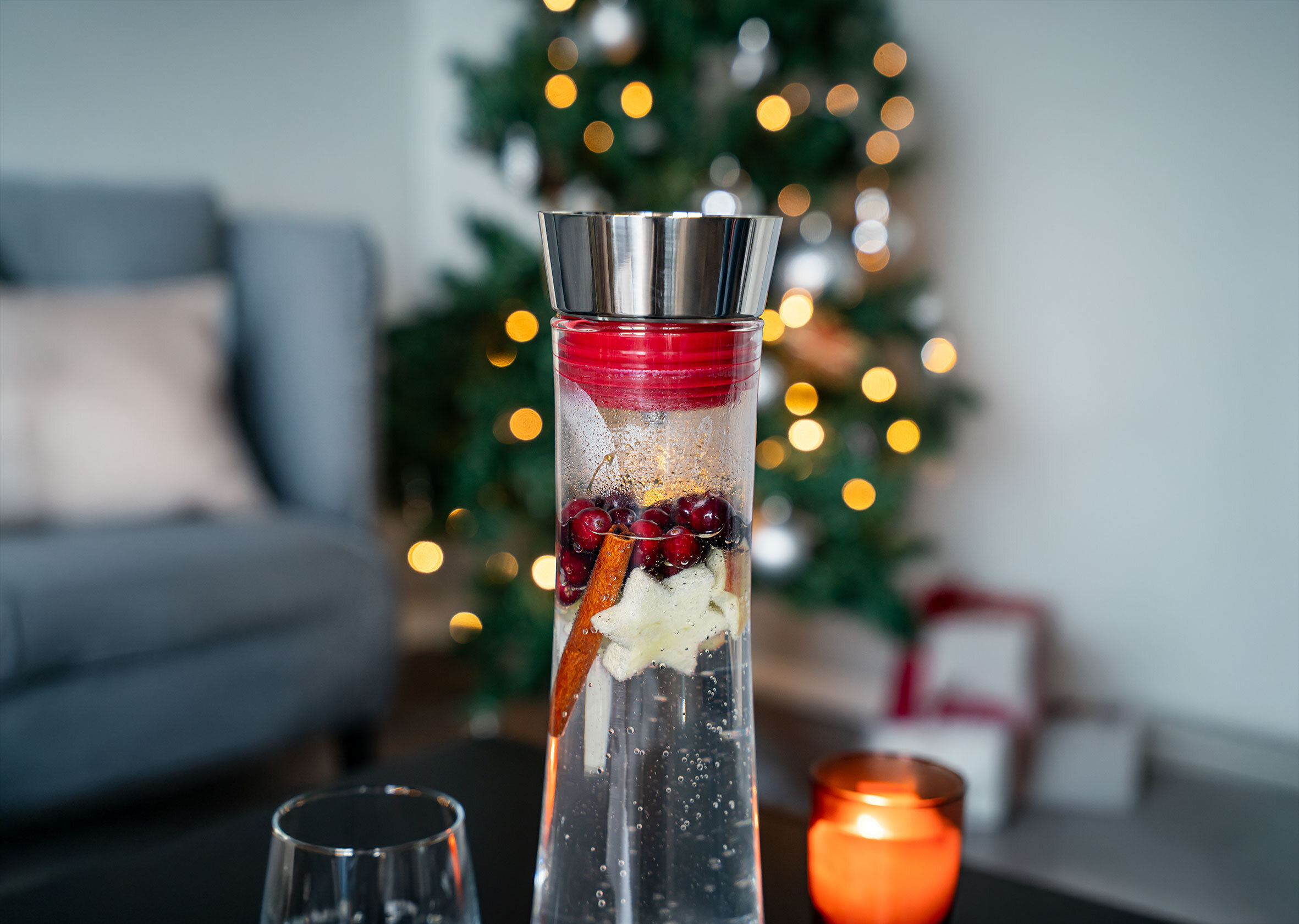 Weihnachtsdeko mit Cranberry-Infusion in einer Glasflasche, vor Weihnachtsbaum und Kissen.