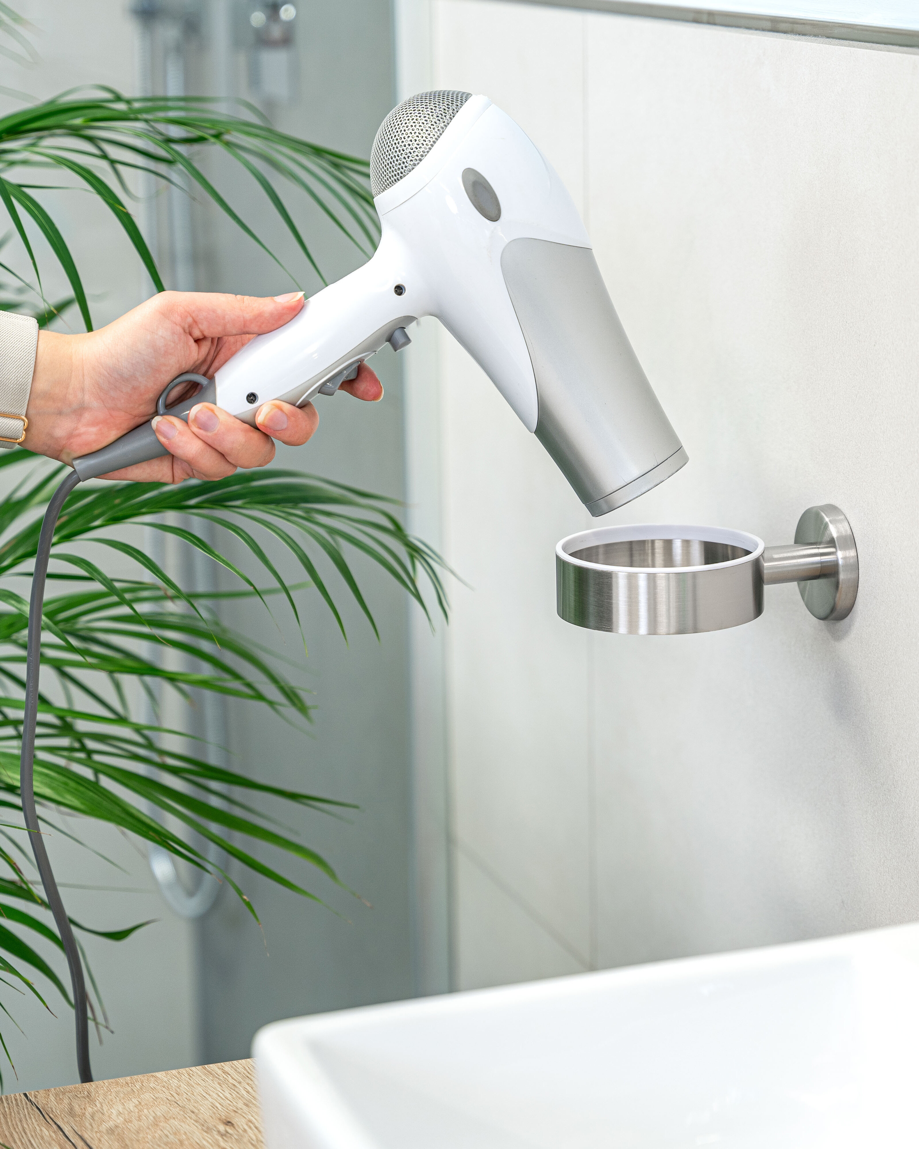 Eine Person steckt einen weißen Haartrockner in eine Wandhalterung in einem modernen Badezimmer. Grüne Pflanzenblätter und ein weißes Waschbecken schaffen eine frische, saubere Atmosphäre.
