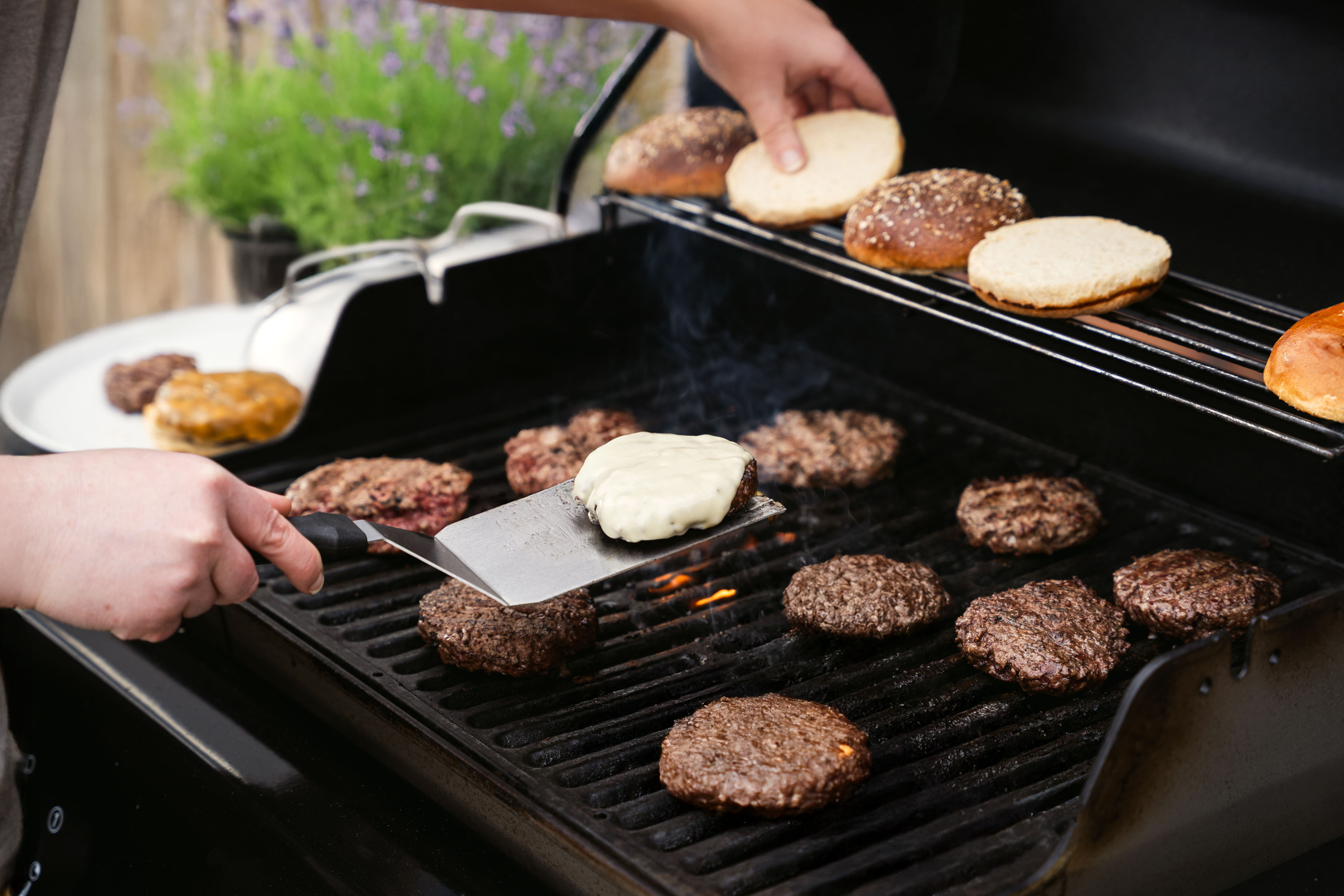 Eine Hand hält einen Spatel und wendet einen Burger mit geschmolzenem Käse auf einem Grill. Im Hintergrund liegen mehrere Burgerbrötchen und gegrillte Rinderpatties.