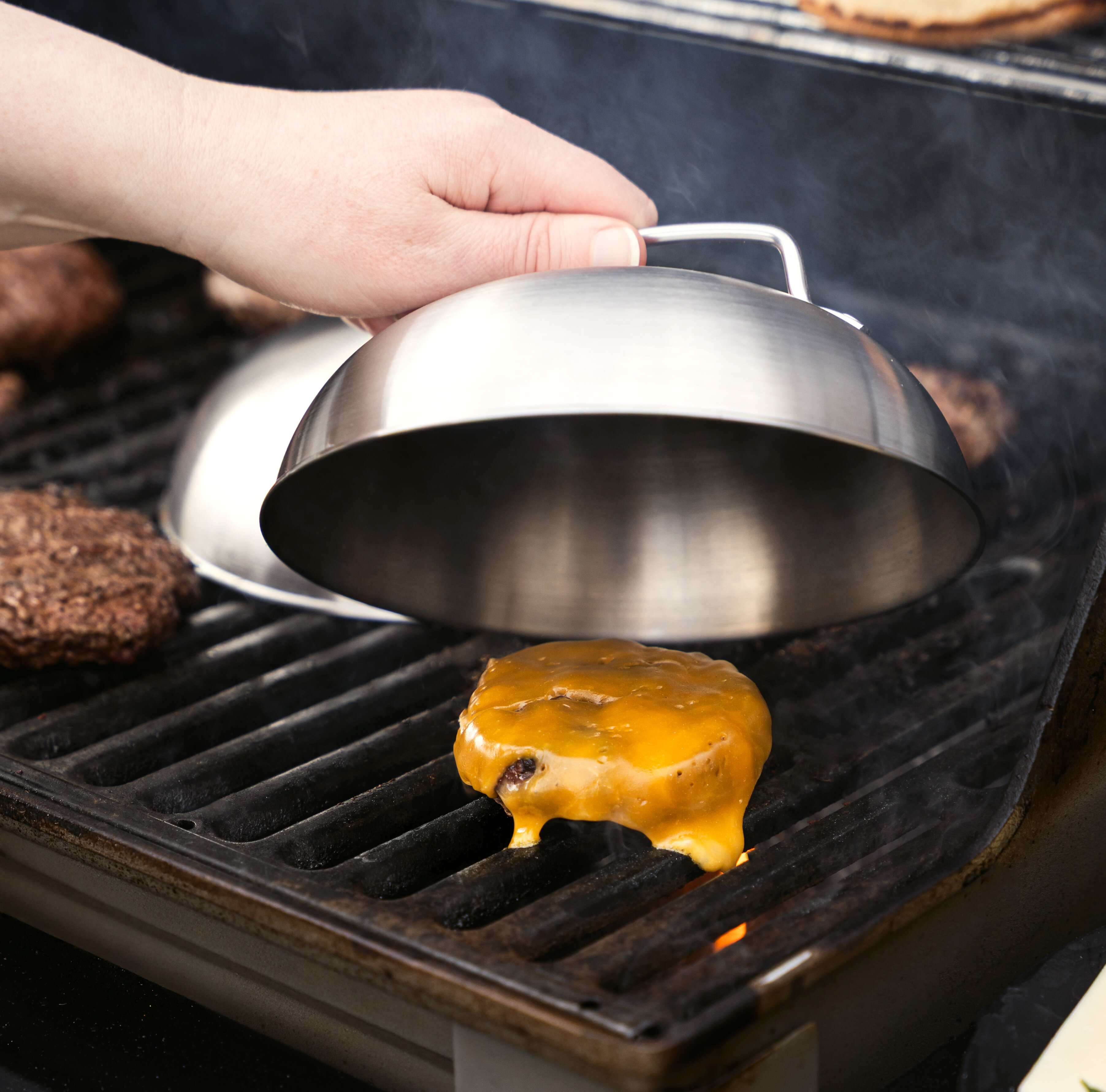 Eine Hand hebt einen silbernen Käse-Schmelzdeckel über einen Burger mit geschmolzenem Käse auf einem Grill. Im Hintergrund sind weitere Burger-Patties sichtbar.