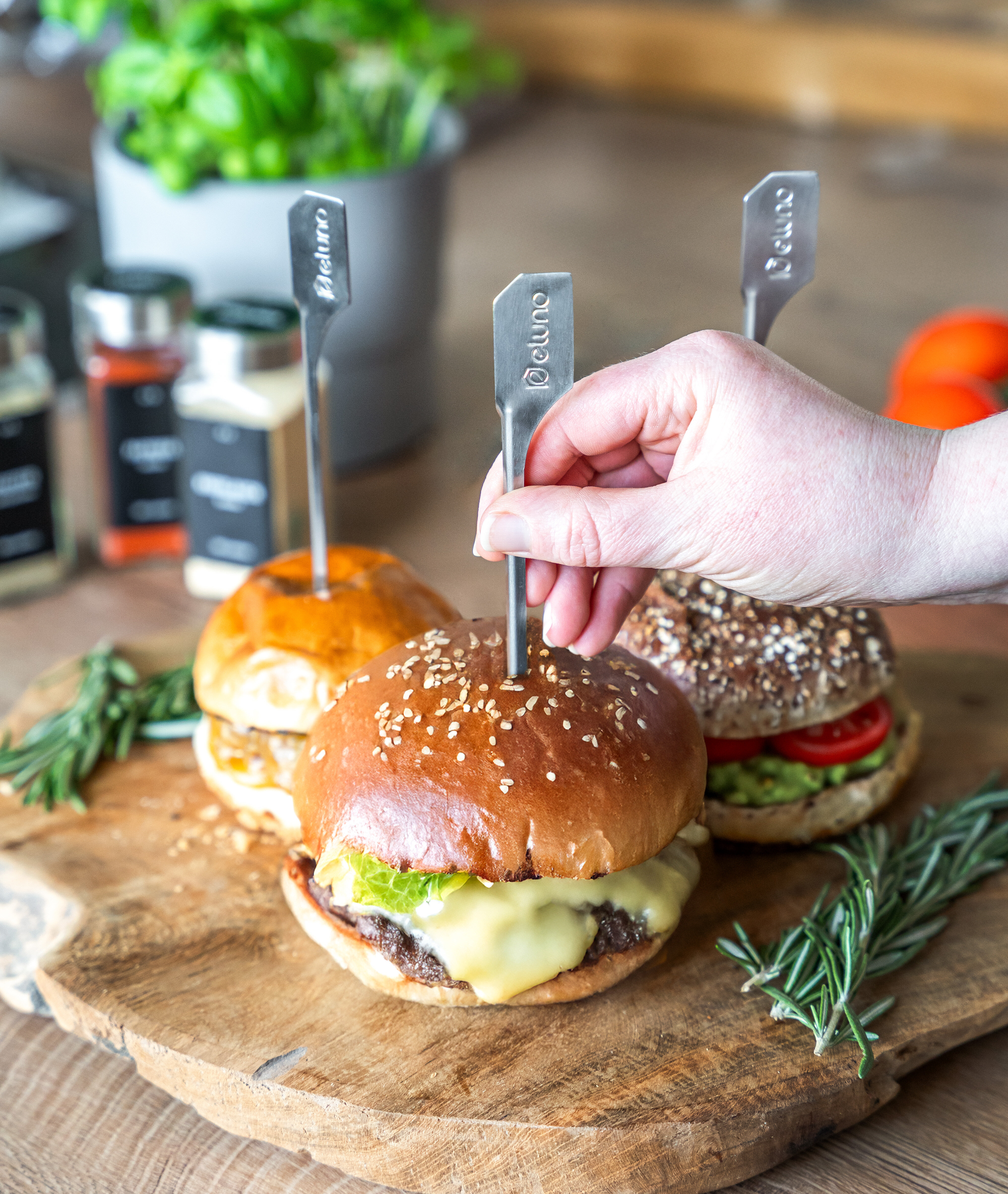 Drei Burger auf einem Holzbrett, einer mit Käse und Salat, einer mit Tomate, einer mit Sesambrötchen. Eine Hand hält einen Burger mit einem Metallspieß.
