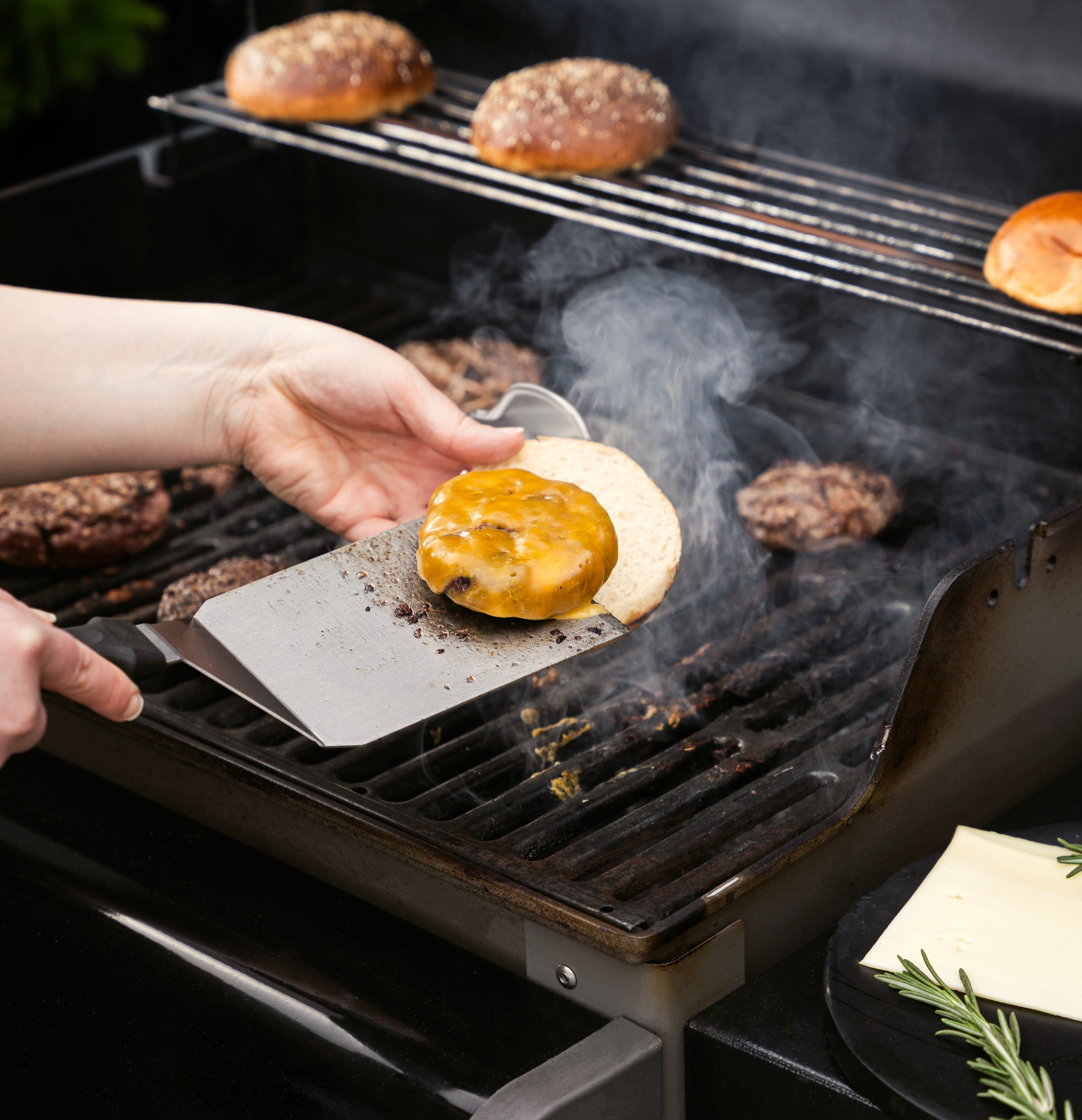 Eine Hand hält einen Spatel mit einem Burger-Patty und geschmolzenem Käse, während sich im Hintergrund weitere Burger-Patties und Brötchen auf einem Grill befinden. Rauch steigt auf.