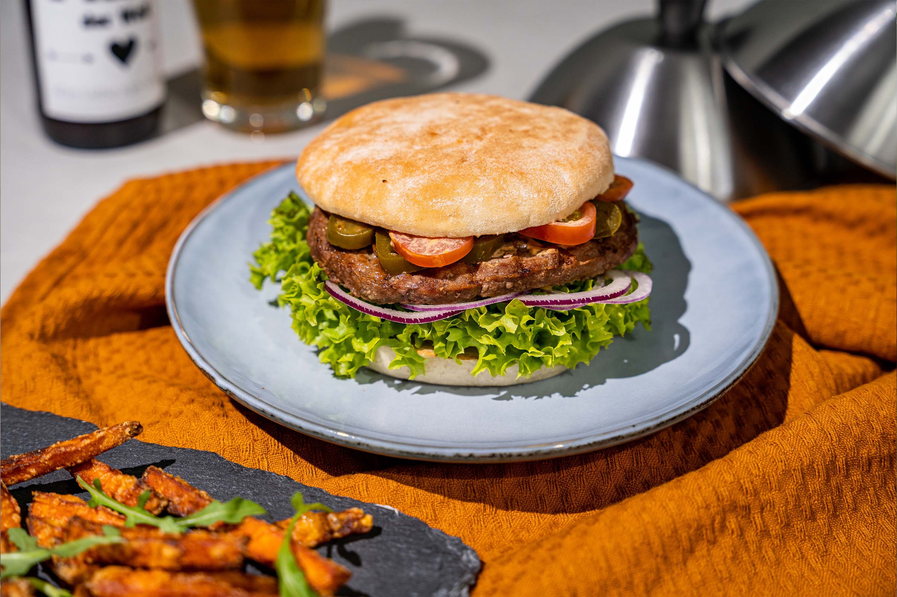 Ein Burger auf einem blauen Teller, belegt mit Salat, Tomaten, Zwiebeln und Gurken, auf einem rustikalen Tuch. Im Hintergrund sind ein Glas Bier und eine Flasche sichtbar.