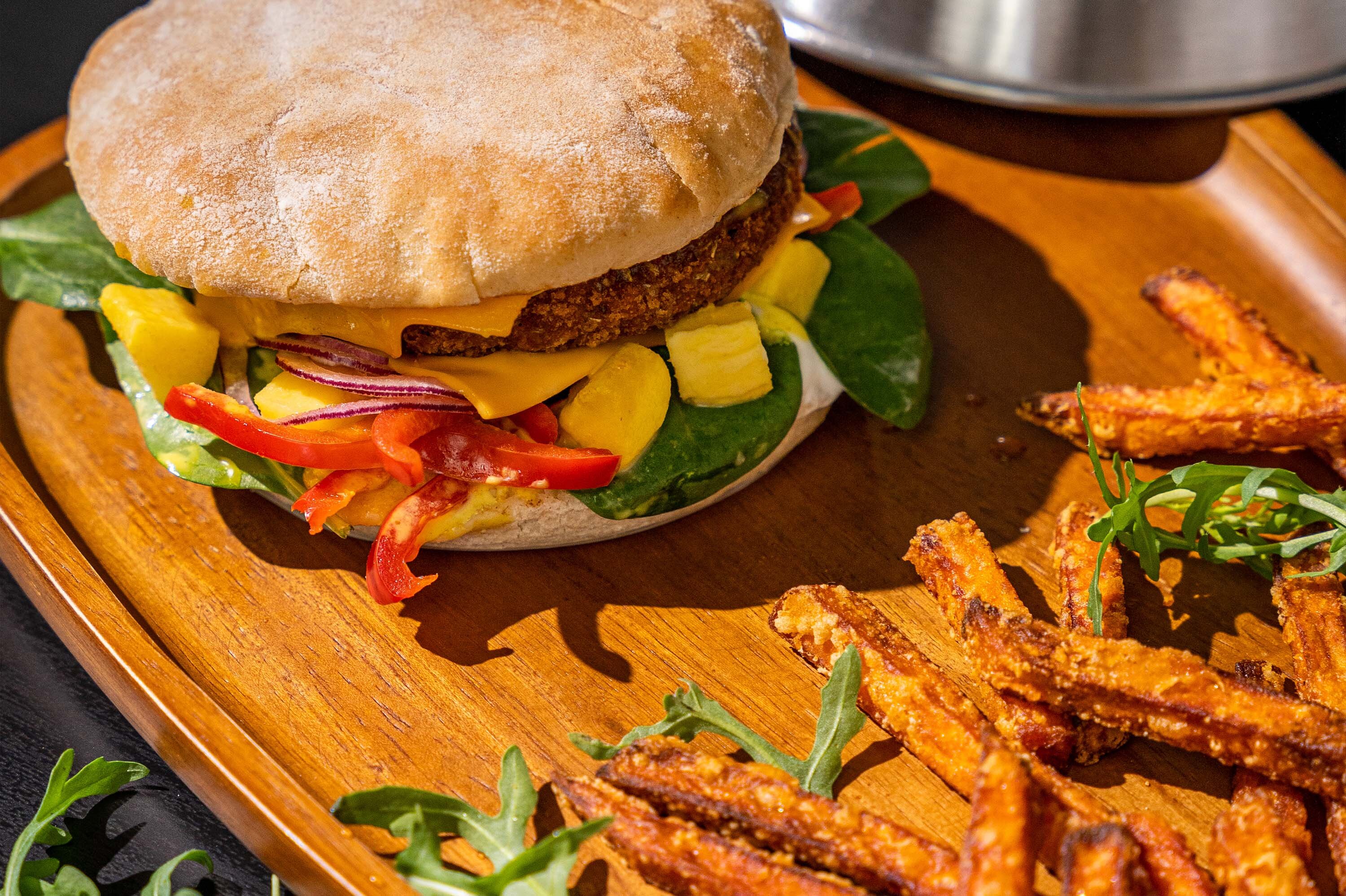 Ein Burger mit einem Brötchen, gefüllt mit einem pflanzlichen Patty, Cheddar-Käse, Spinat, Paprika, Mango und Zwiebeln. Daneben liegt eine Portion Süßkartoffelpommes auf einem Holztablett.