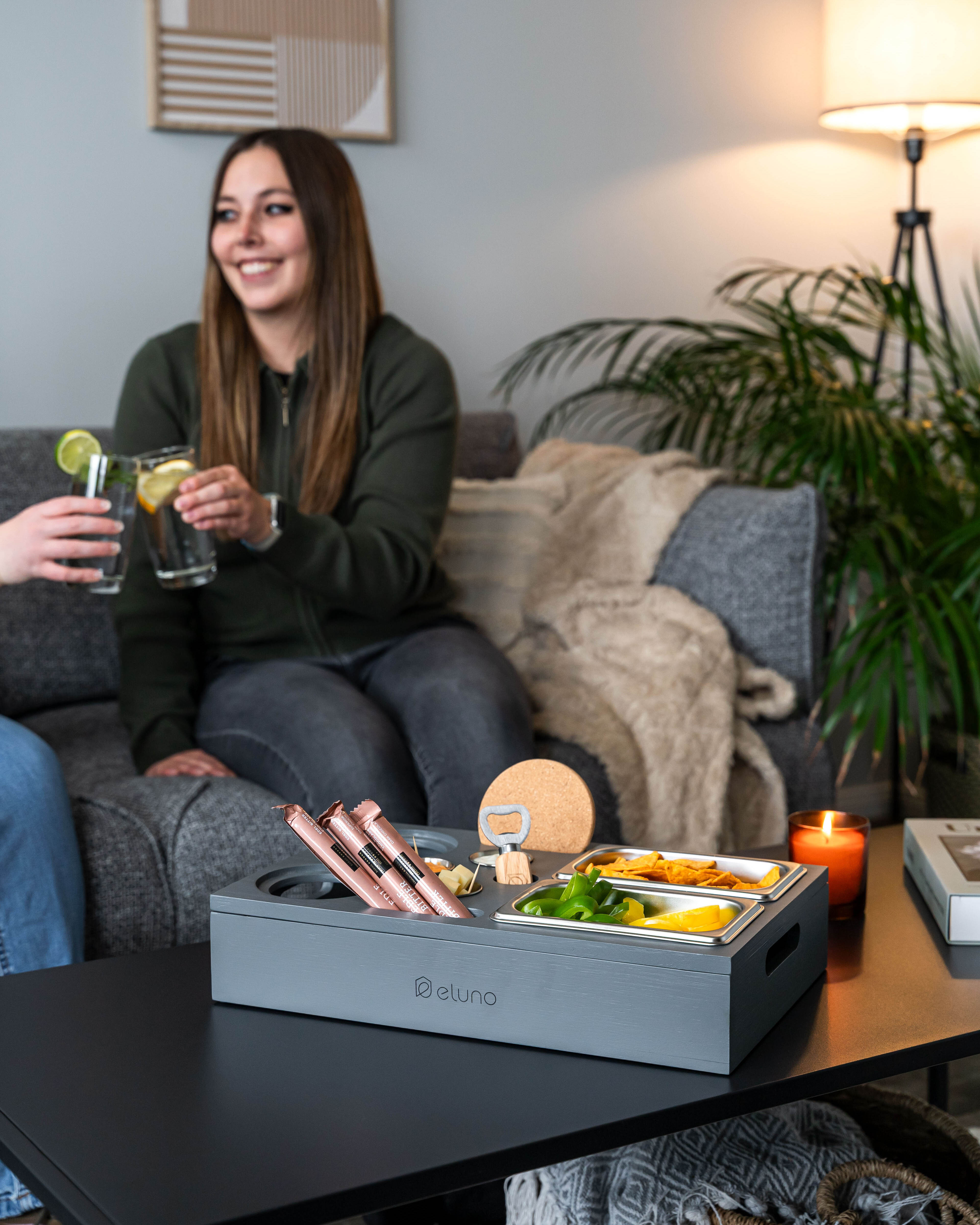 Graue Couchbar mit Fächern für Snacks und Getränke auf schwarzem Tisch, im Hintergrund graues Sofa mit Decke und Pflanze.