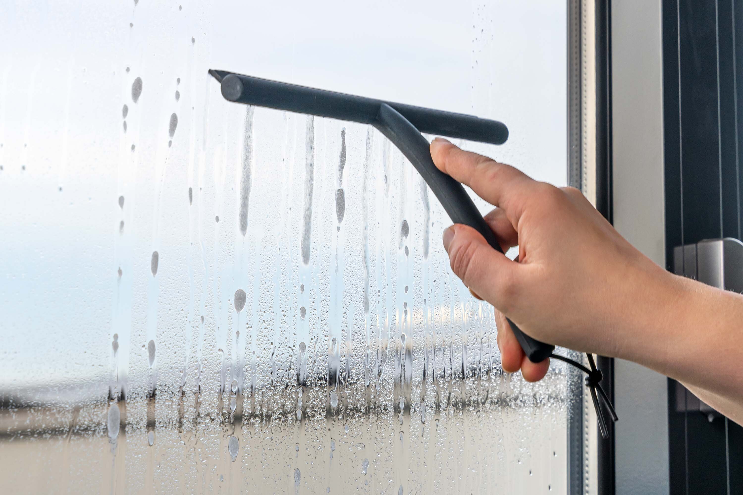 Eine Hand hält einen Duschabzieher mit schwarzem Griff, der auf einer beschlagenen Fensterscheibe verwendet wird. Wassertröpfchen sind sichtbar.