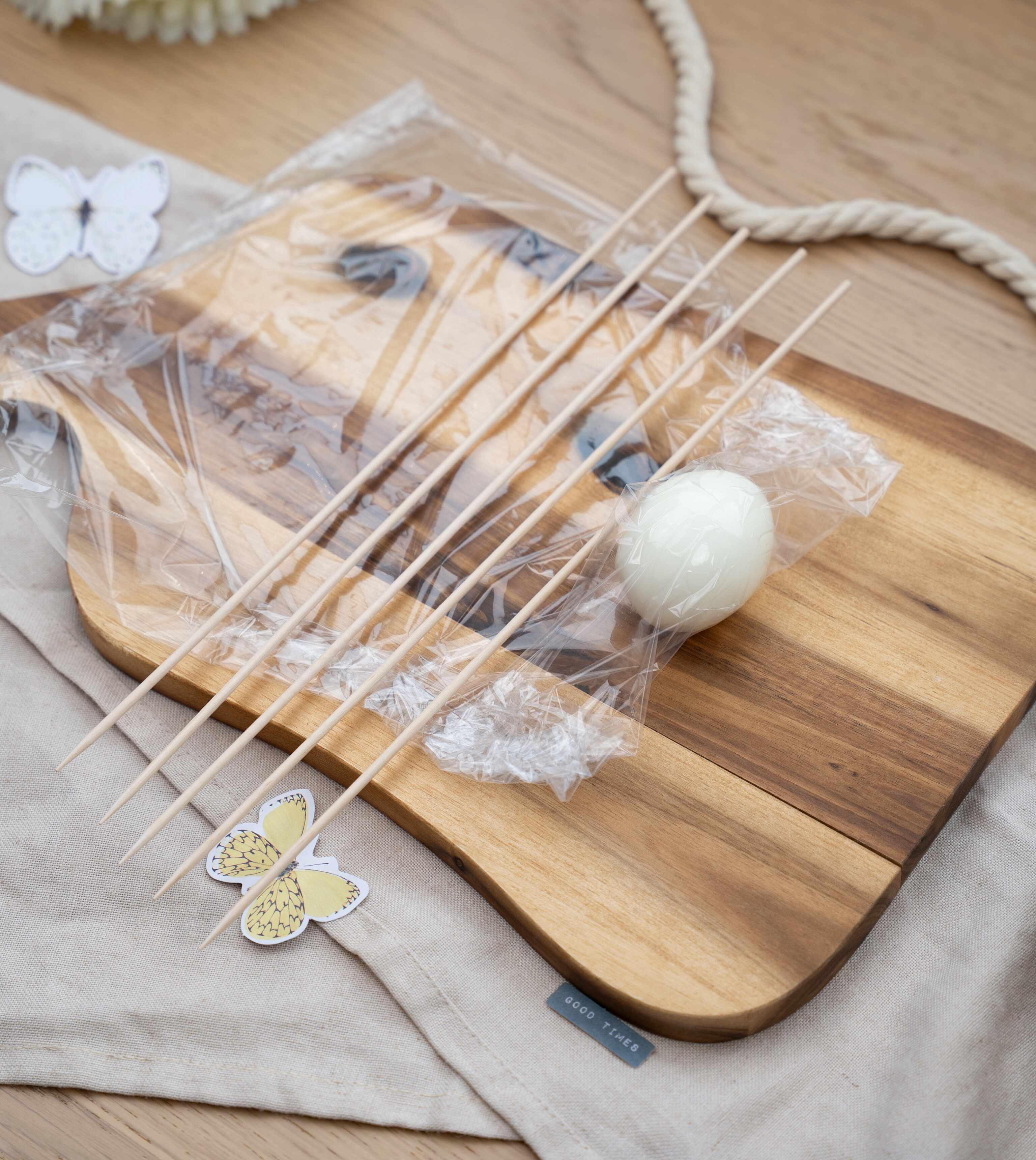 Holzbrett mit mehreren Holzspießen, einer verpackten weißen Kugel und Papier-Schmetterlingen auf einem hellen Stoff