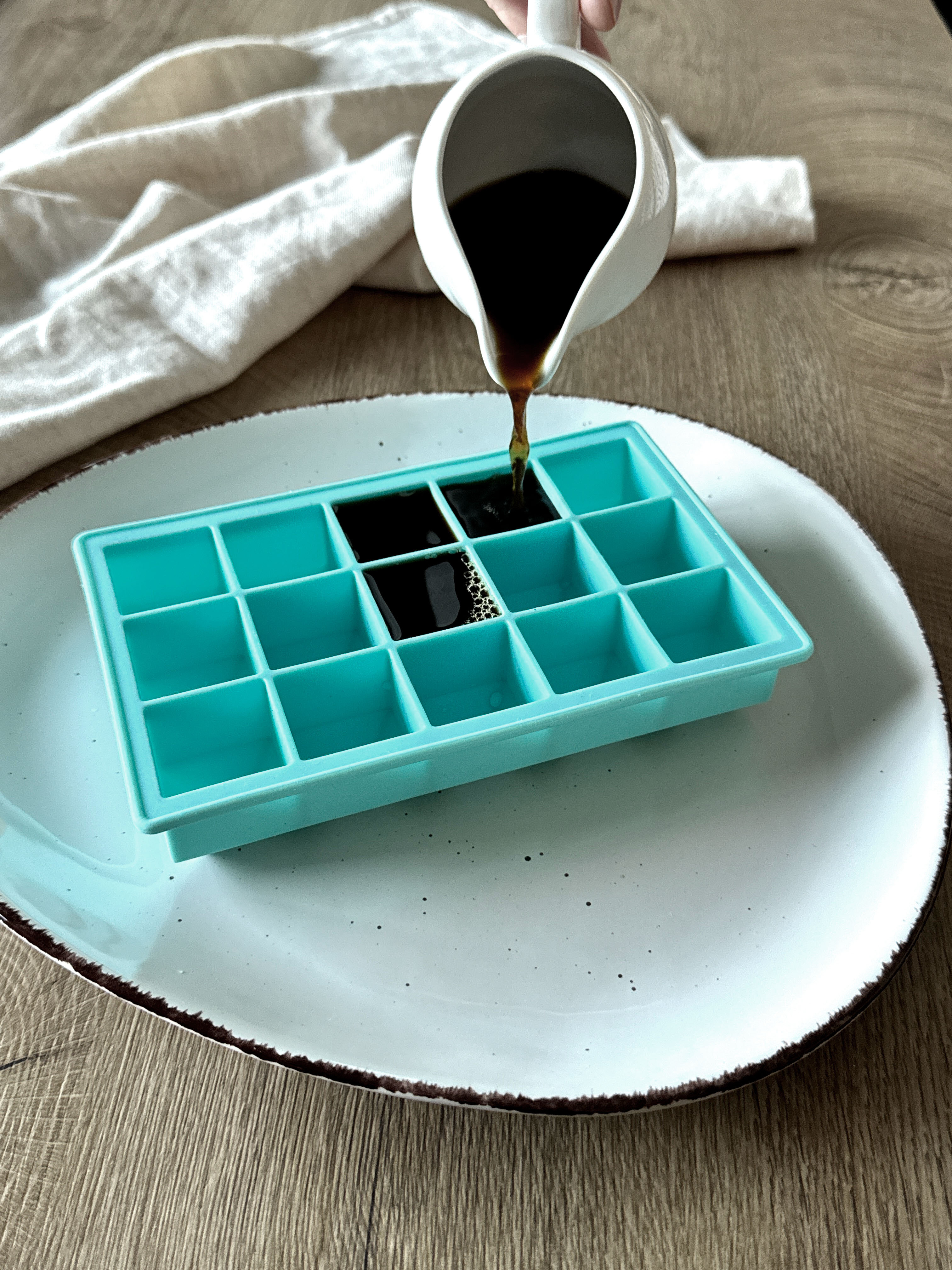 Eine Hand gießt eine dunkle Flüssigkeit aus einer weißen Kanne in eine türkisfarbene Silikonform mit 16 Fächern auf einem weißen Teller.