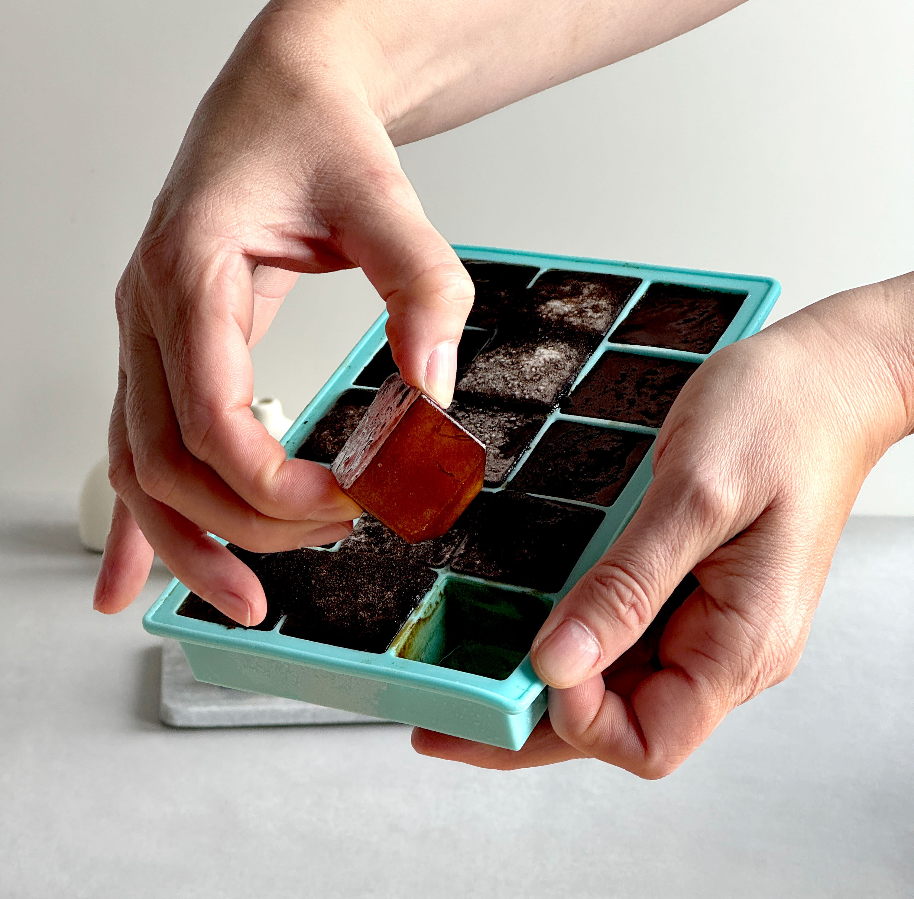 Eine Person hält einen Eiswürfel aus einer blauen Silikonform mit 16 Fächern. Die Hand ist in der Nähe eines braunen Eiswürfels, während die andere Hand die Form hält.