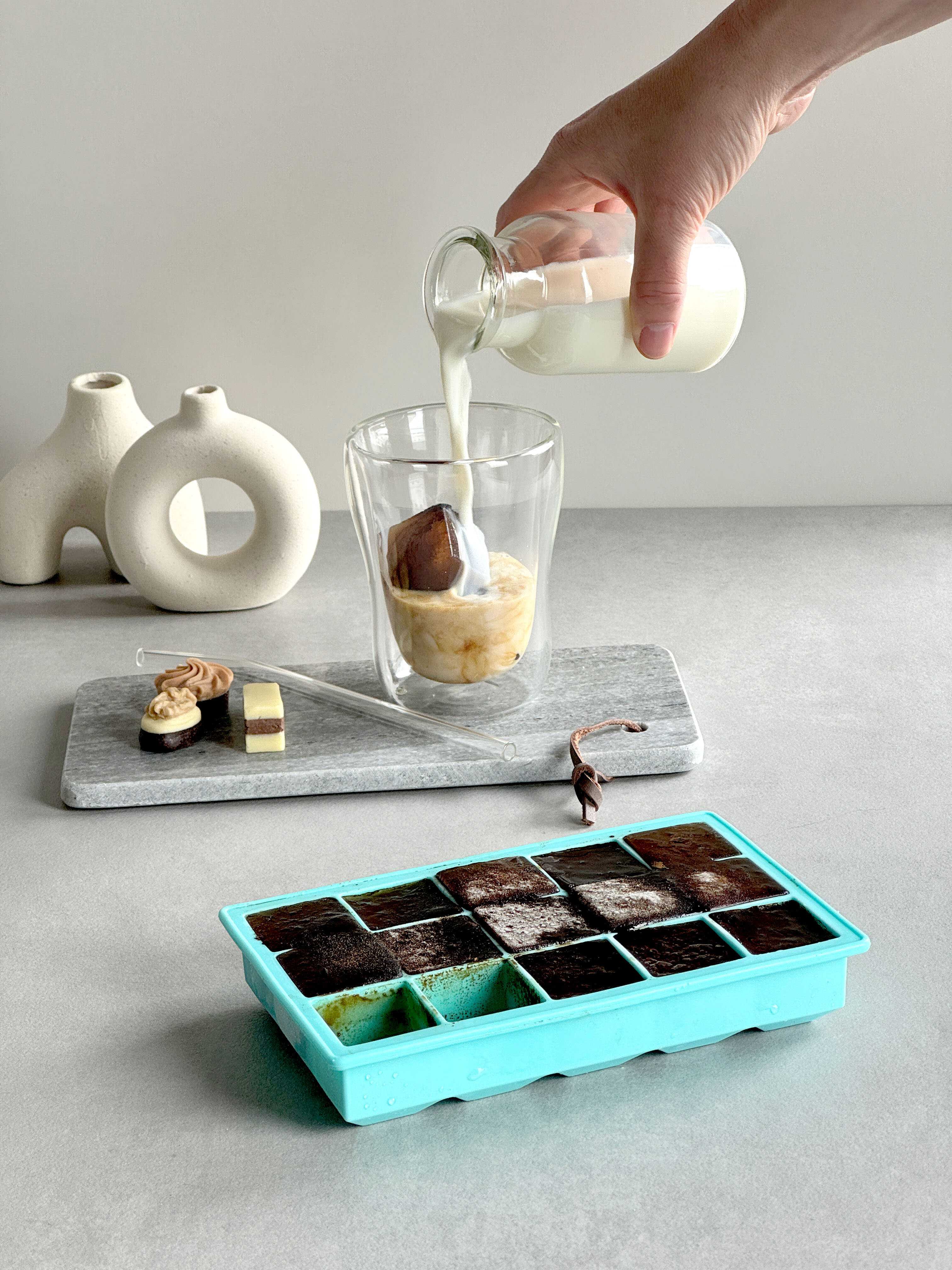 Eine Hand gießt Milch in ein Glas mit Eiswürfeln und einer Schokoladensorte. Im Vordergrund liegt eine Eiswürfelschale in Türkis mit gefrorenem Kaffee.