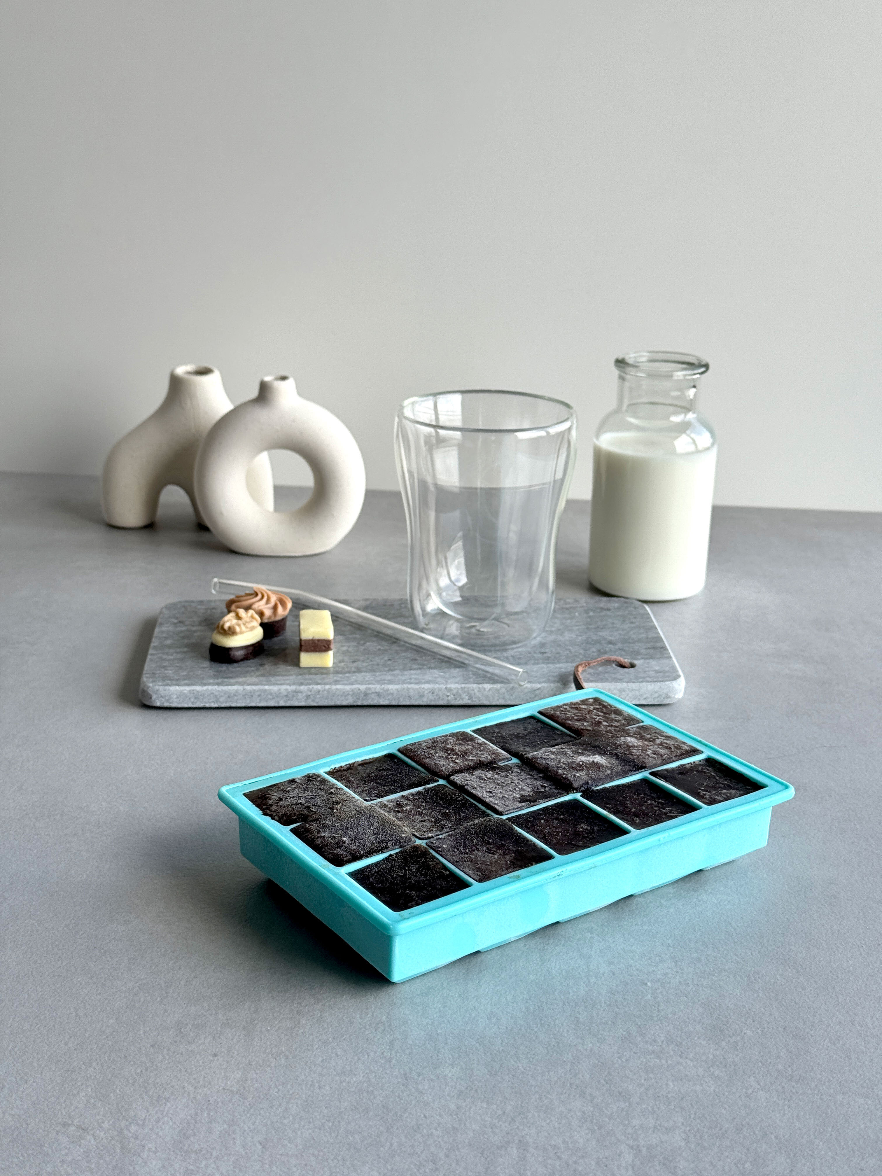 Eine türkisfarbene Eiswürfelschale mit 16 Fächern steht auf einem grauen Tisch. Im Hintergrund sind ein Glas, eine Glasflasche mit Milch, ein Teller mit Süßigkeiten und zwei dekorative Vasen zu sehen.