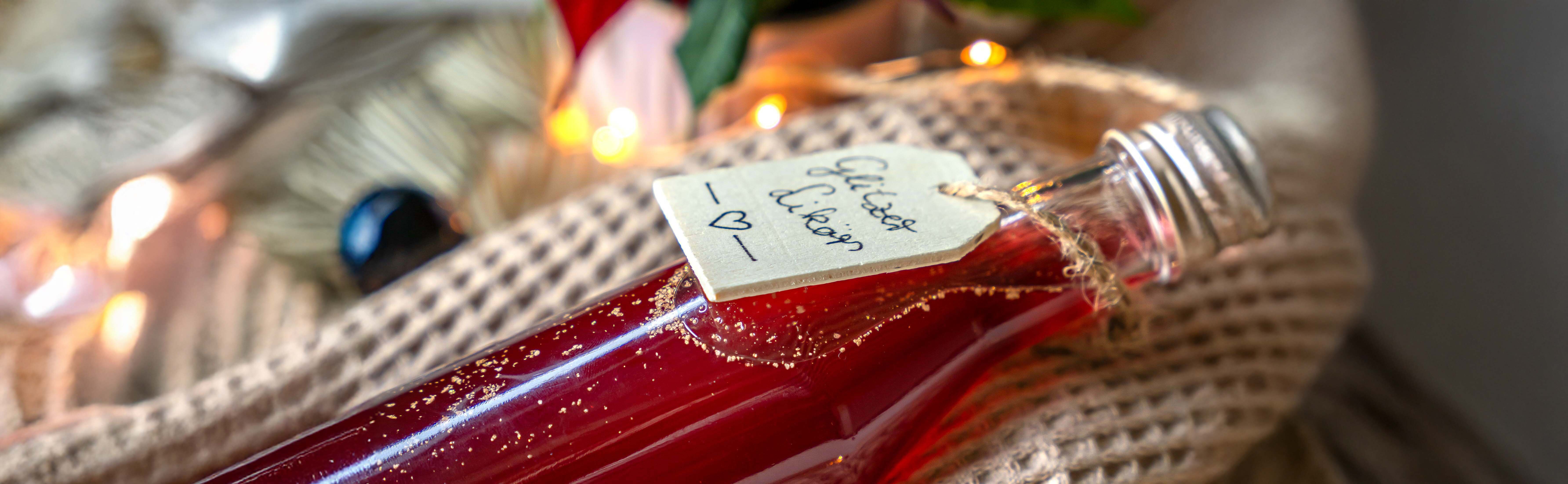 Schlanke Glasflasche mit rotem Glitzerlikör, mit handbeschriftetem Anhänger 'Glitzer Likör' auf beigem Strickstoff liegend.