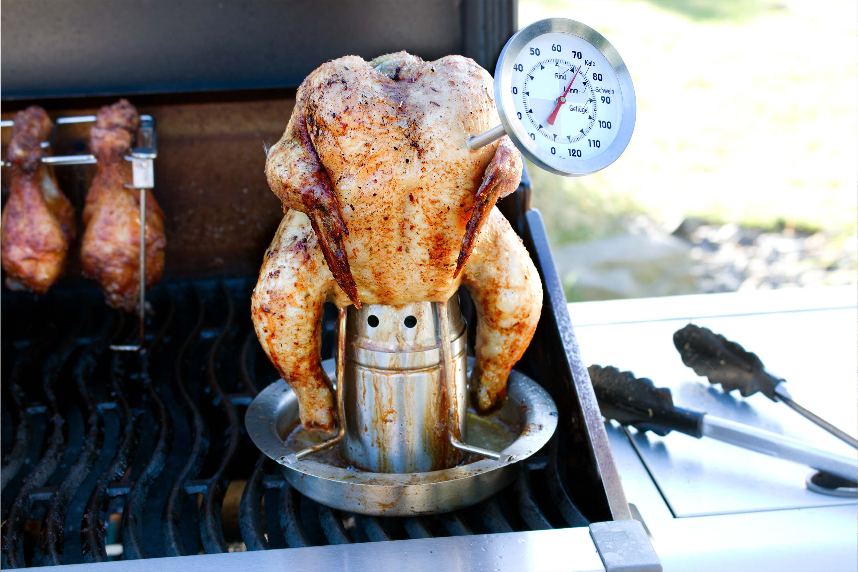 Ein gegrilltes Hähnchen auf einem Grill, aufrecht auf einem speziellen Halter mit einem Thermometer. Das Hähnchen ist gewürzt und hat eine goldbraune Farbe.