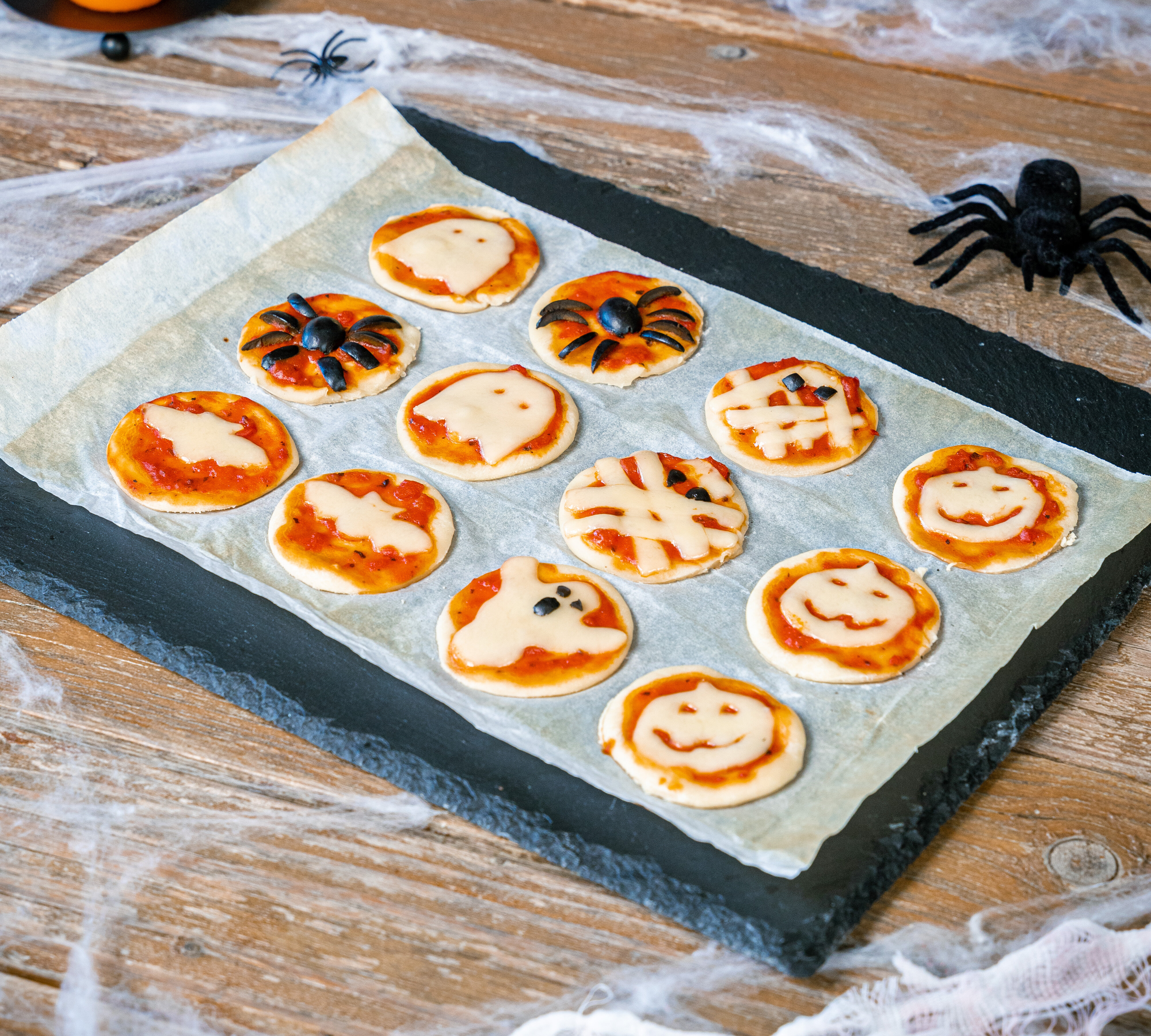 Ein Blech mit 12 kleinen Halloween-Pizzen in verschiedenen Formen, darunter Spinnen, Kürbisse und Gespenster, mit Käse und Tomatensauce.