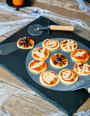 Runde Pizzastücke mit Halloween-Motiven wie Spinnen, Gespenstern und Fledermäusen auf einem Pizzaschneider. Im Hintergrund sind Spinnweben und eine orangefarbene Kerze.