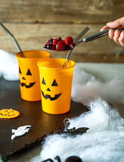 Zwei orangefarbene Becher mit Halloween-Gesicht, gefüllt mit einer Mischung aus Beeren. Eine Hand hält einen Löffel mit Beeren über den Bechern. Im Hintergrund sind Dekorationen und Spinnweben.