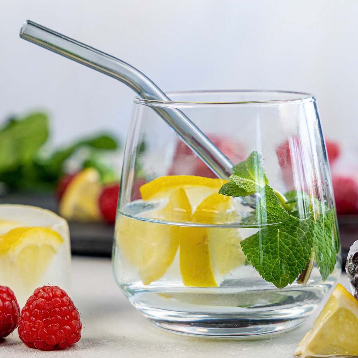 Ein Glas mit Wasser, Zitronenscheiben und frischer Minze, ausgestattet mit einem Metallstrohhalm. Im Vordergrund sind Himbeeren und Zitronenscheiben sichtbar.