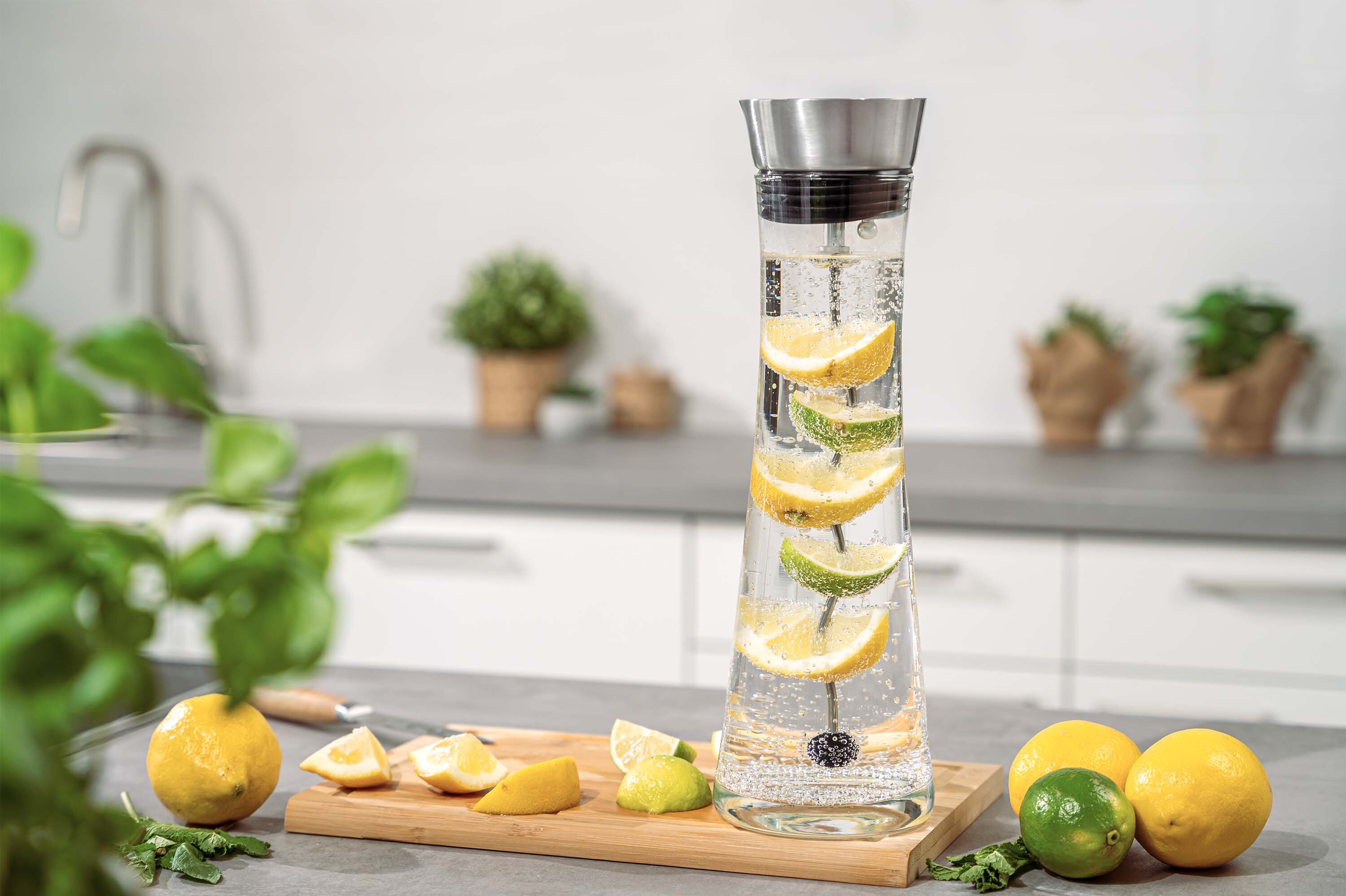 Ein Glasbehälter mit Zitronen- und Limettenscheiben, gefüllt mit sprudelndem Wasser, steht auf einem Holzbrett. Im Hintergrund sind Pflanzen und eine moderne Küchenzeile sichtbar.