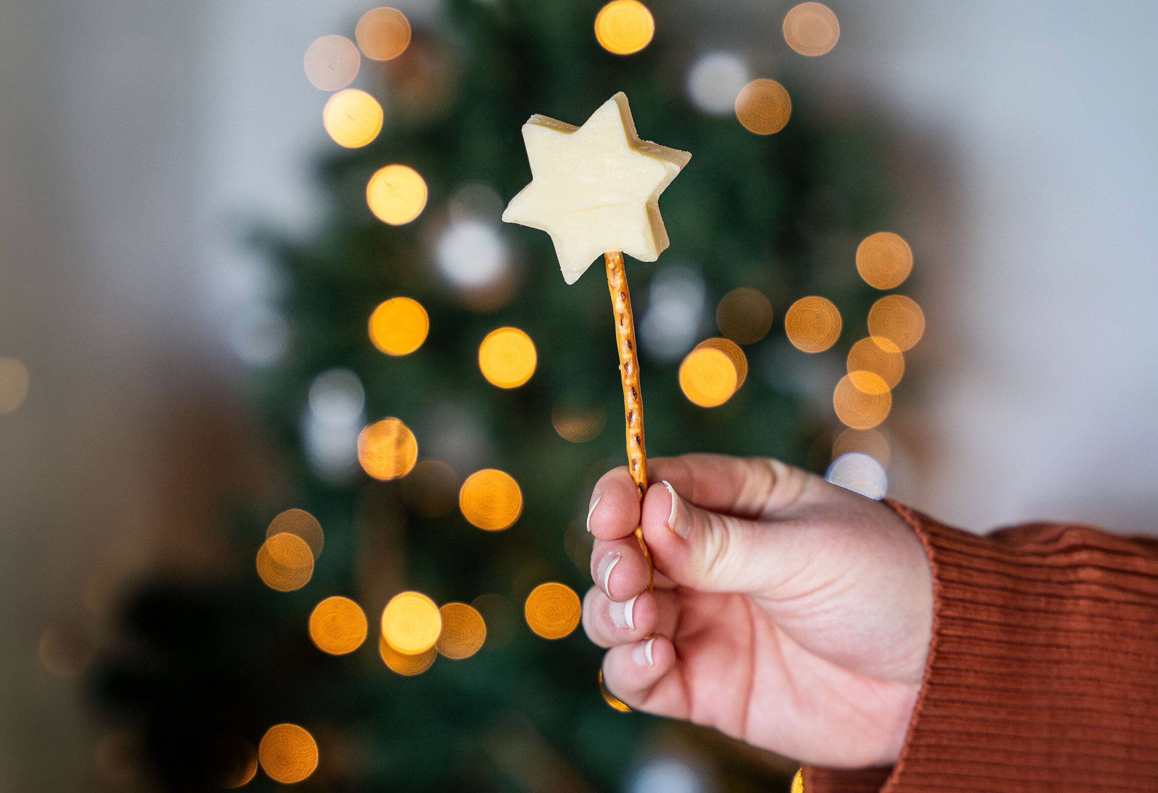 Hand hält einen Stern-förmigen Keks auf einem dünnen Stab vor unscharfem Weihnachtsbaum mit Lichterketten.