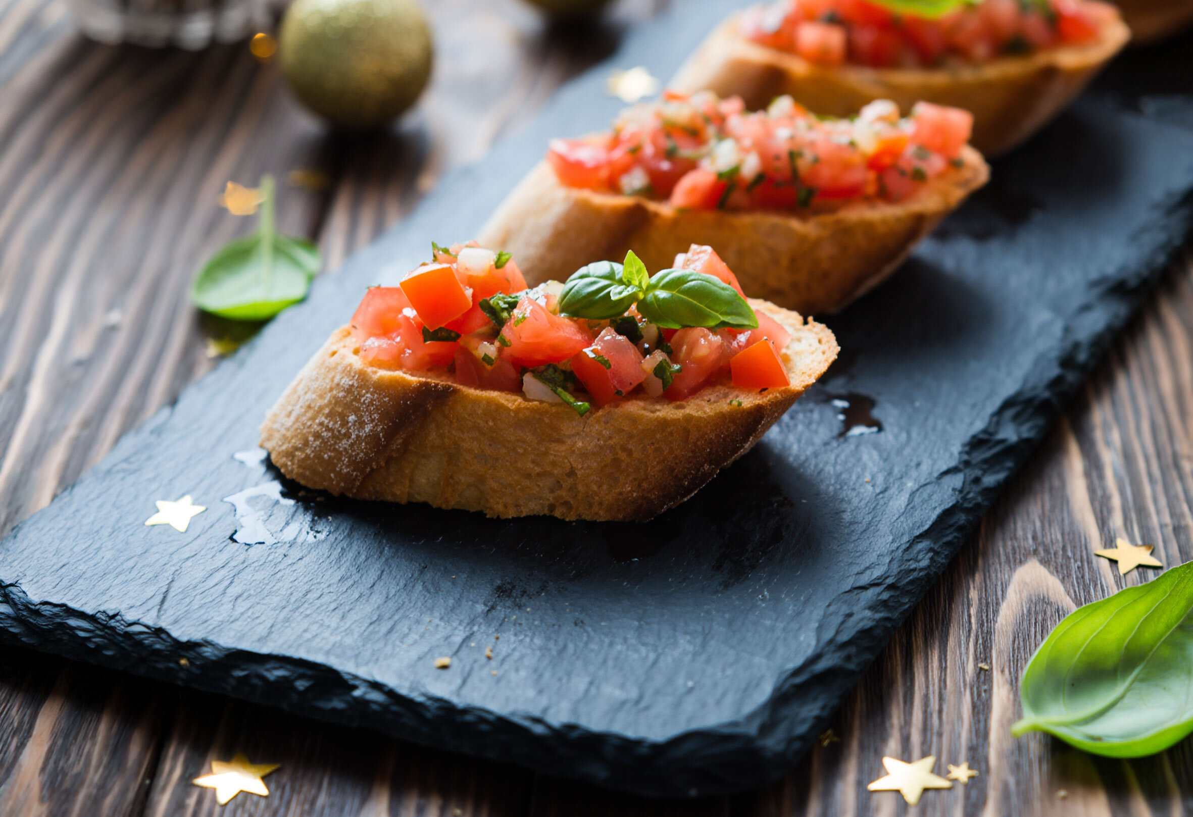Drei Bruschetta mit gewürfelten Tomaten und Basilikum auf dunkler Schieferplatte, umgeben von grünen Basilikumblättern und goldenen Sternen.