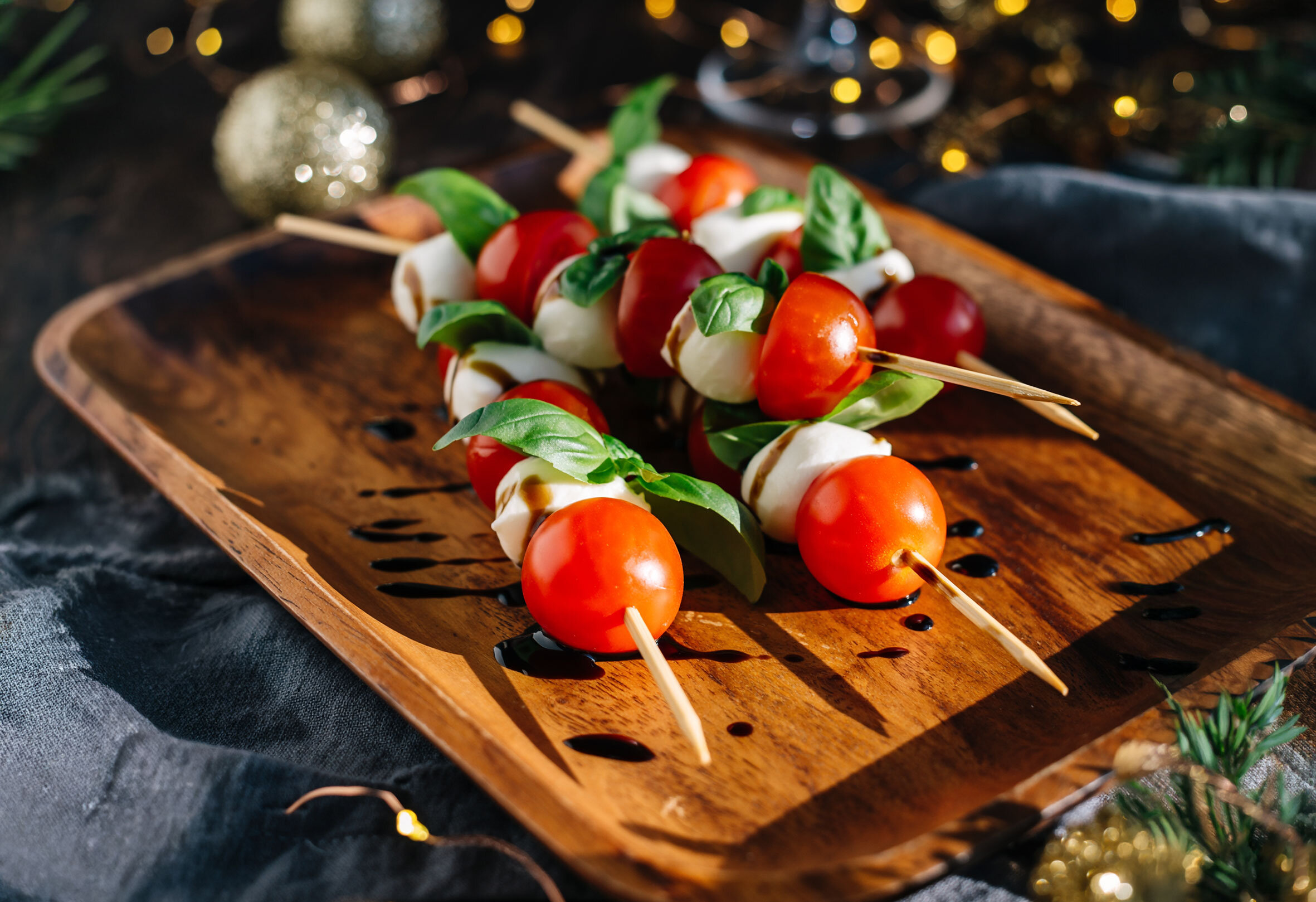 Drei Caprese-Spieße mit Kirschtomaten, Mozzarellakugeln und Basilikumblättern auf einem Holzbrett mit Balsamico-Tropfen.