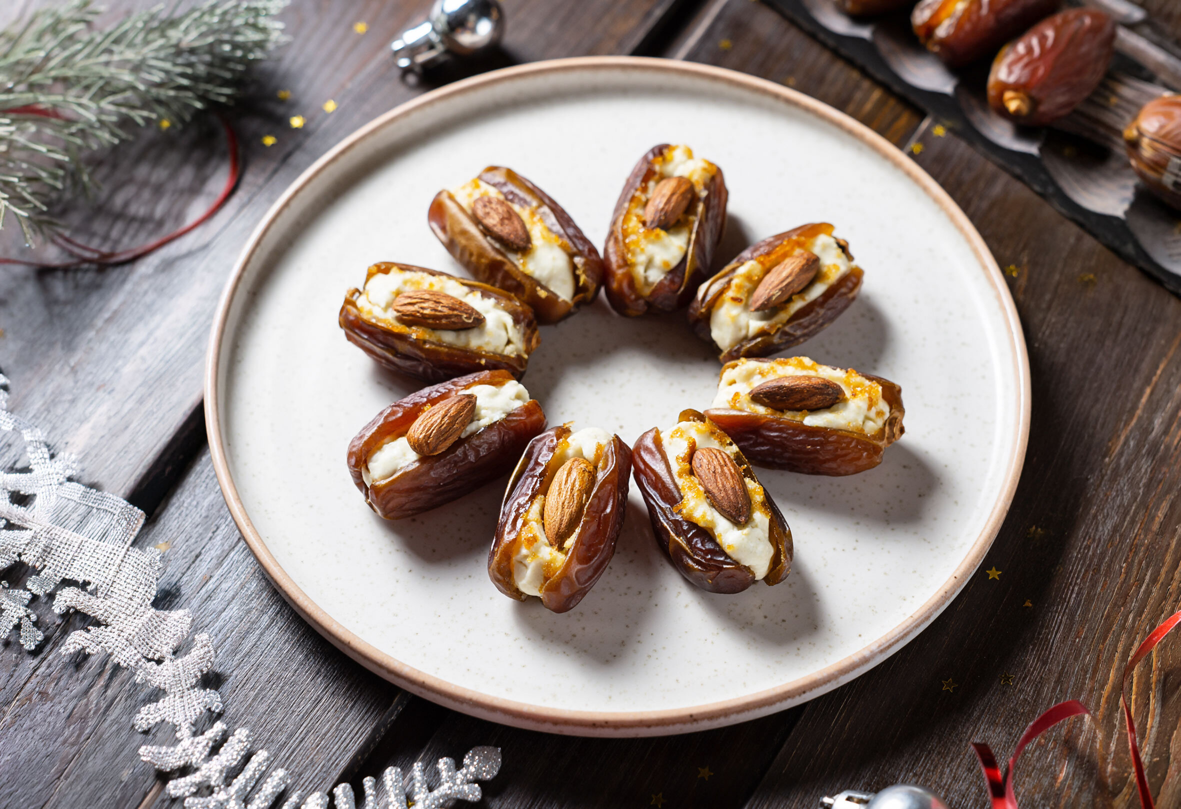 Acht halbierte Datteln auf einem weißen Teller, gefüllt mit Frischkäse und jeweils mit einer Mandel belegt, auf dunklem Holztisch mit Weihnachtsdeko.