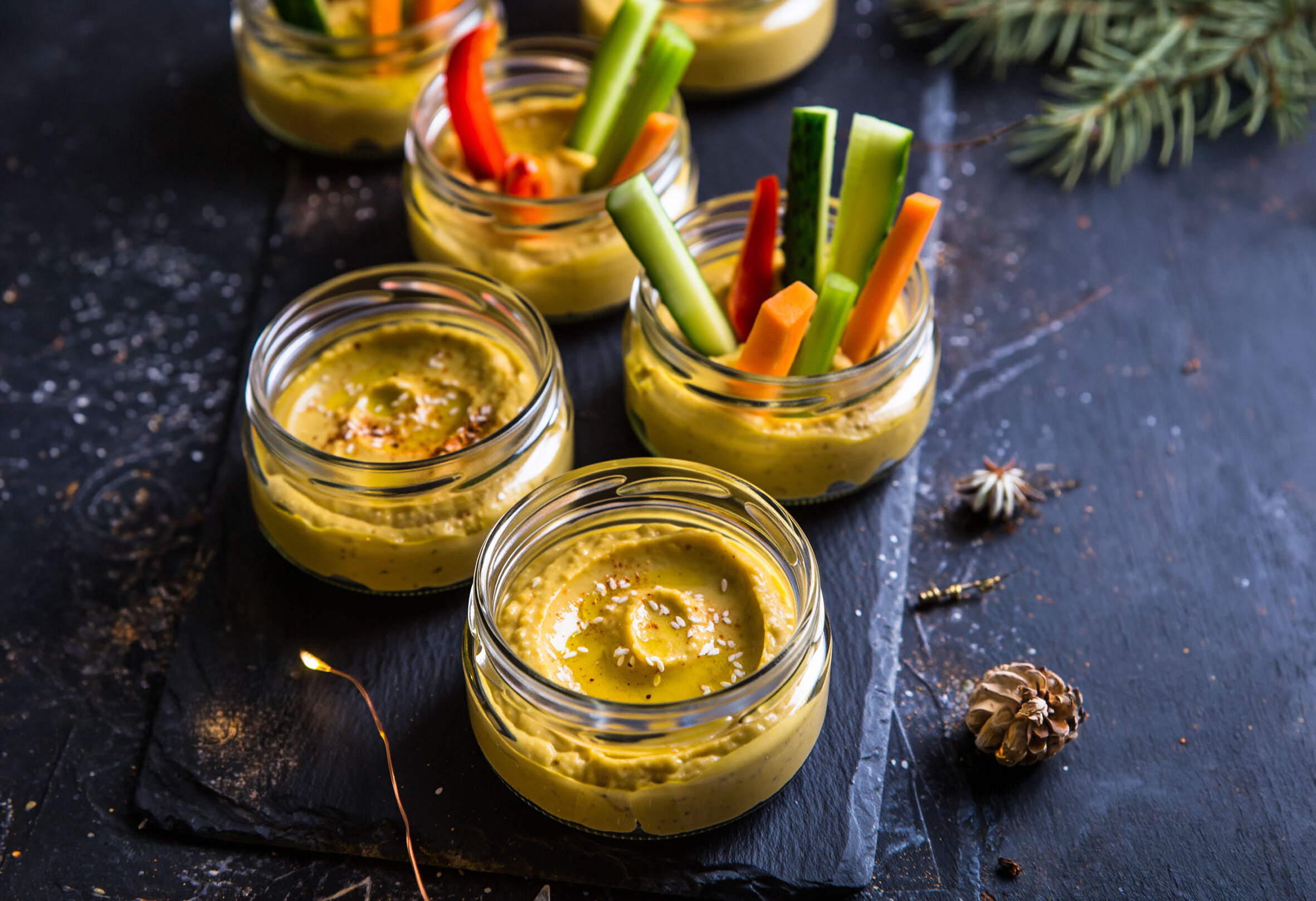 Mehrere kleine Gläser mit gelbem Hummus, eines davon mit Sticks aus Gurke, Karotte und roter Paprika, auf dunklem Untergrund.