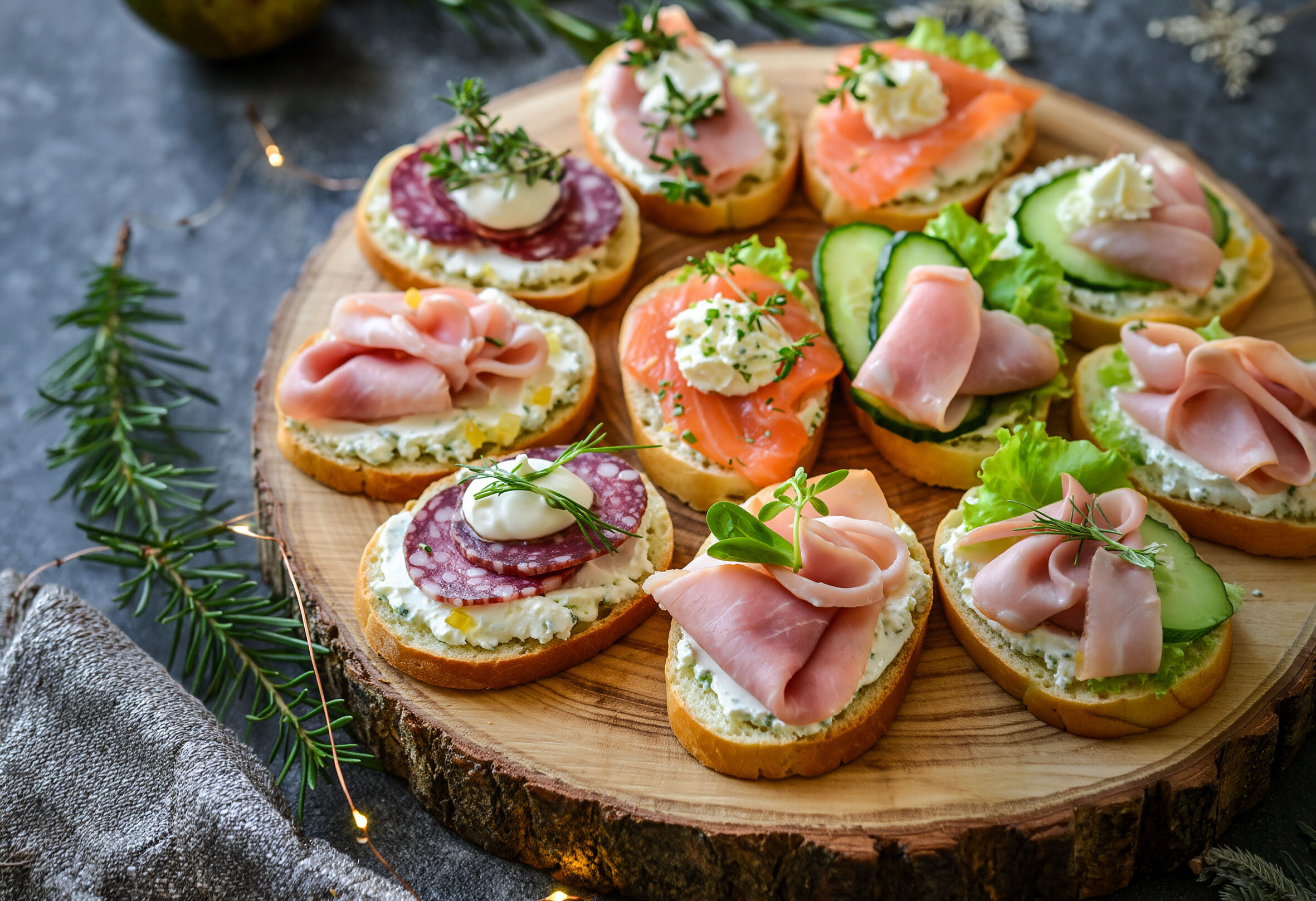 Holzbrett mit verschiedenen belegten Schnittchen, darunter mit Schinken, Salami, Lachs, Gurke und Frischkäse, garniert mit Kräutern.