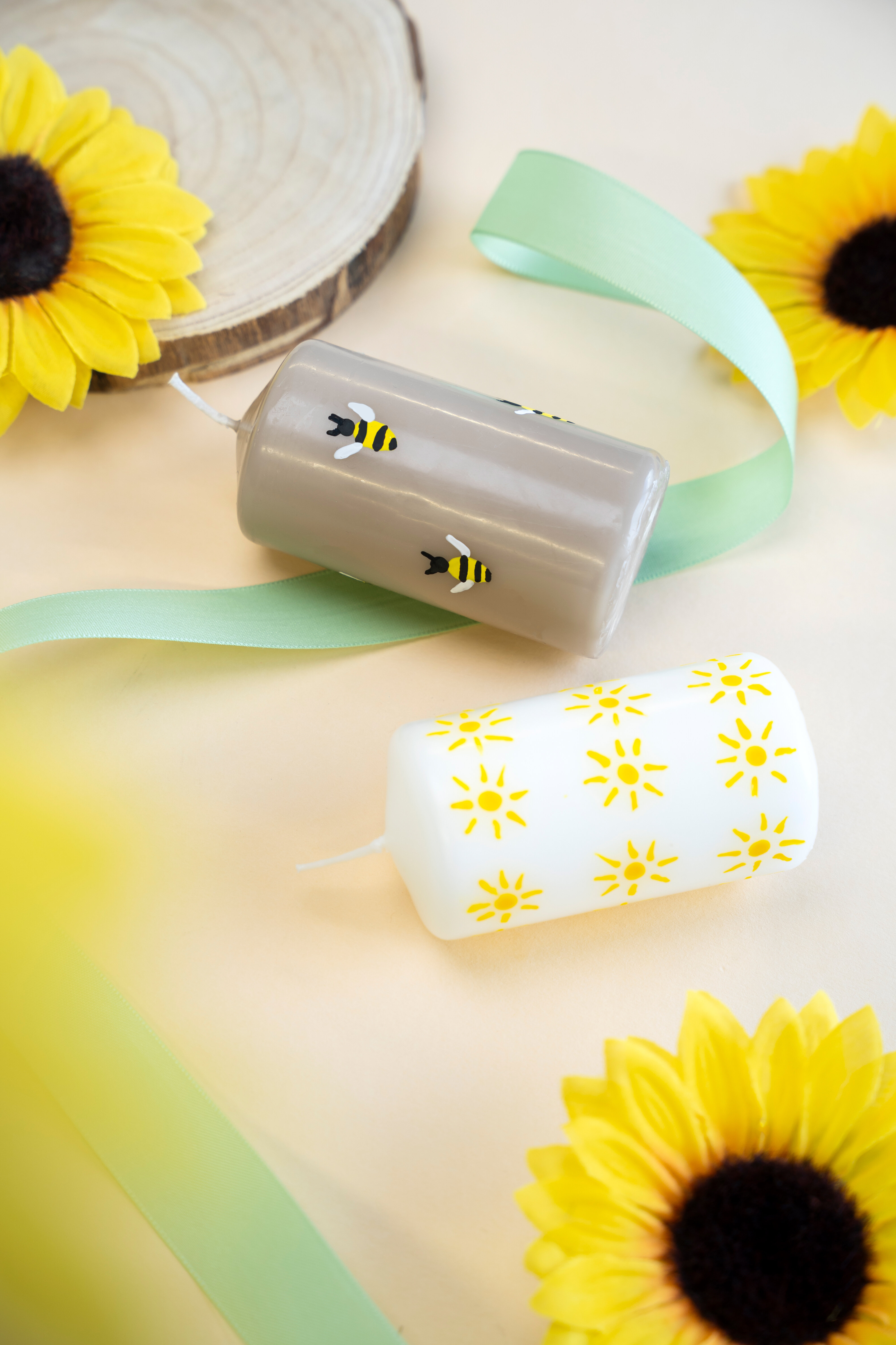 Braune Blockkerze mit Bienen-Motiv bemalt. Darunter eine weiße Blockkerze, die mit Sonnen bemalt wurde.