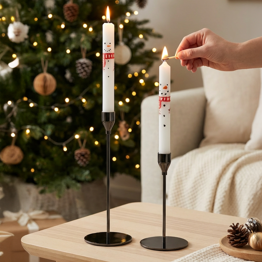 Zwei weiße Kerzen mit Schneemannmotiven auf schwarzen Kerzenhaltern, eine wird mit einem Streichholz angezündet, im Hintergrund ein geschmückter Weihnachtsbaum.