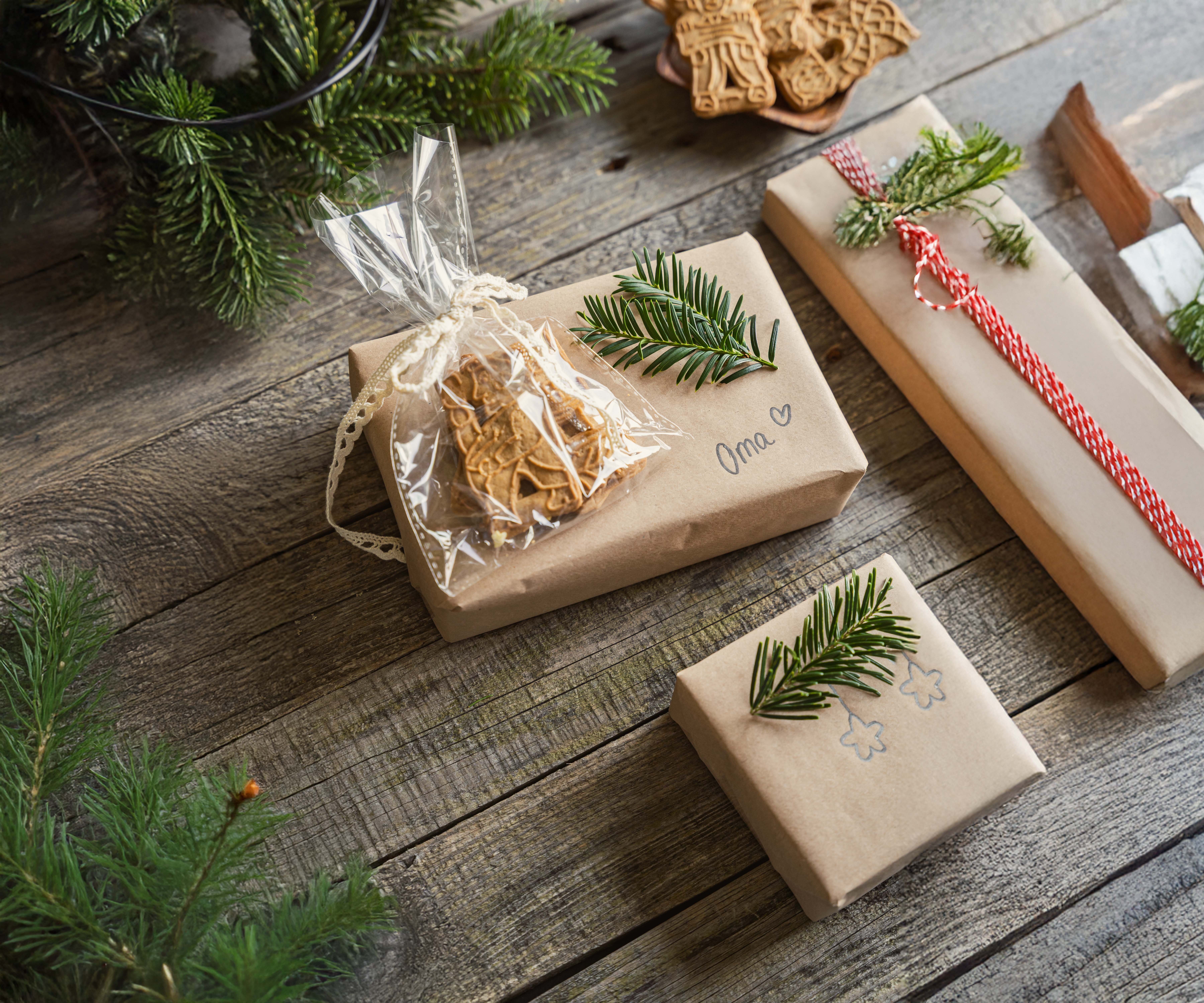 Mehrere mit braunem Papier verpackte Geschenke liegen auf einem Holztisch, dekoriert mit Tannenzweigen, roten Bändern und einem kleinen durchsichtigen Beutel mit Lebkuchen.
