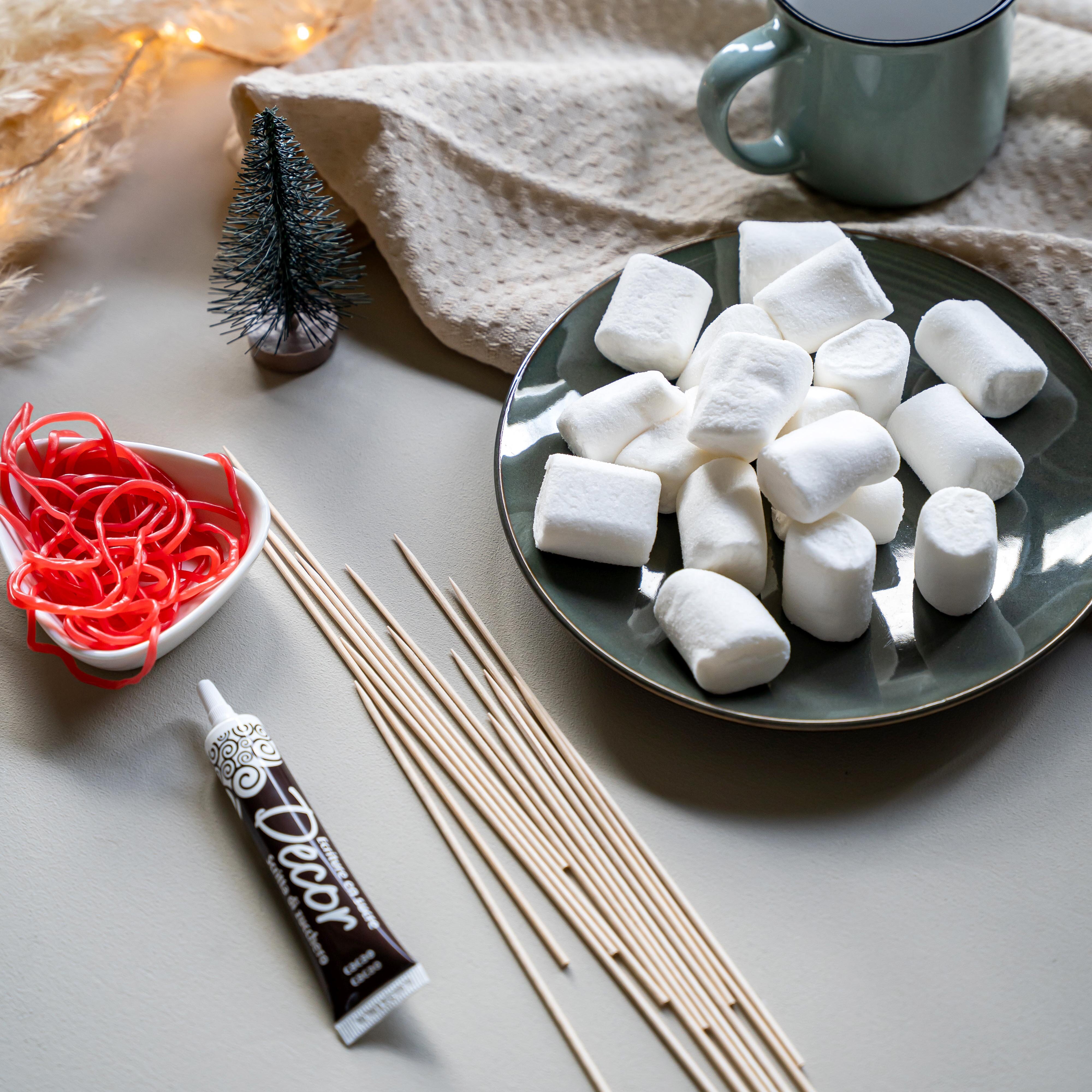Teller mit mehreren weißen Marshmallows, daneben Holzspieße, rote Schnüre in Herzschale, schwarze Dekorcreme-Tube, kleine Tannendekoration und graue Tasse auf Tisch.
