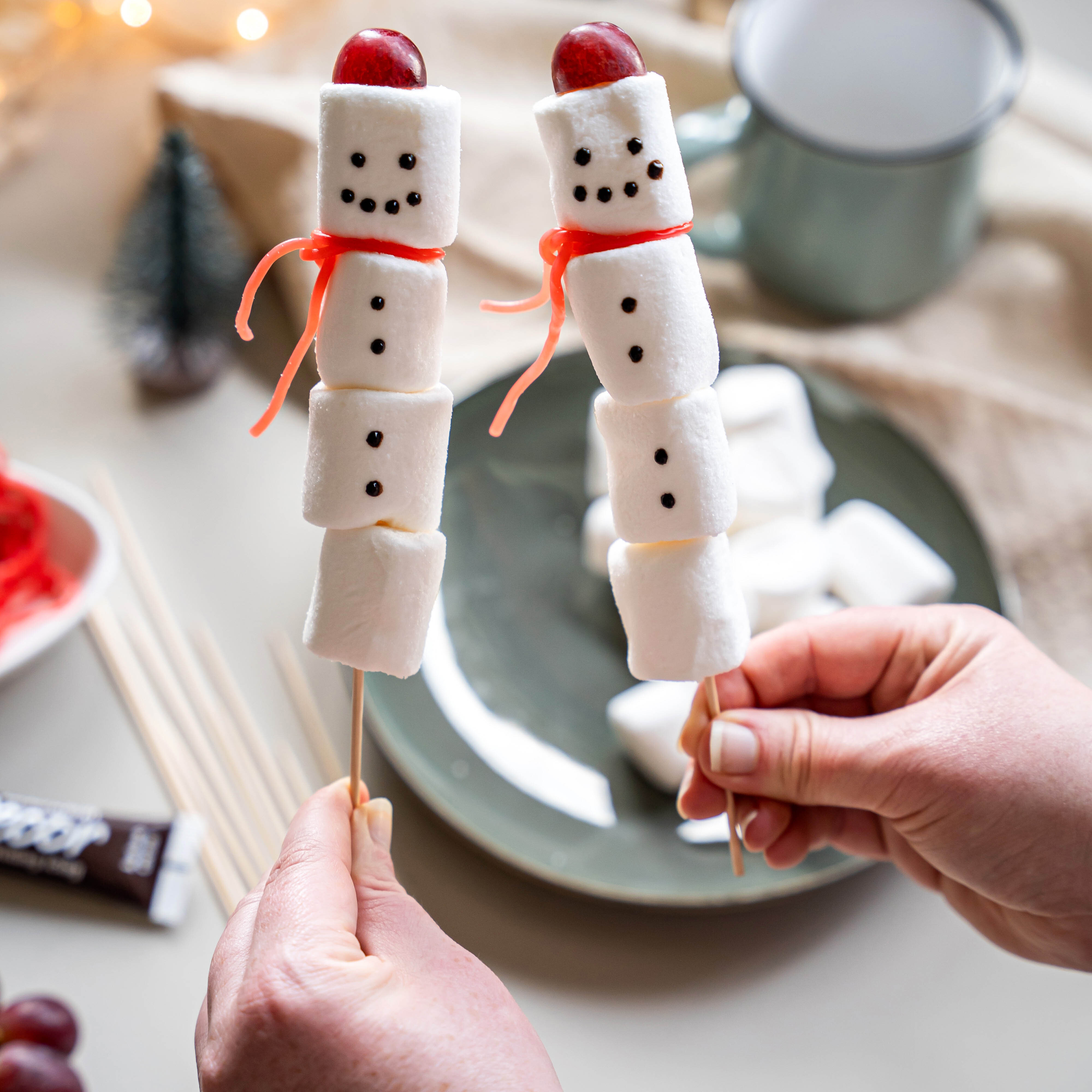 Zwei Schneemänner aus weißen Marshmallows auf Holzspießen, mit roten Schleifen und roten Kirschen als Köpfe, gehalten von Händen.