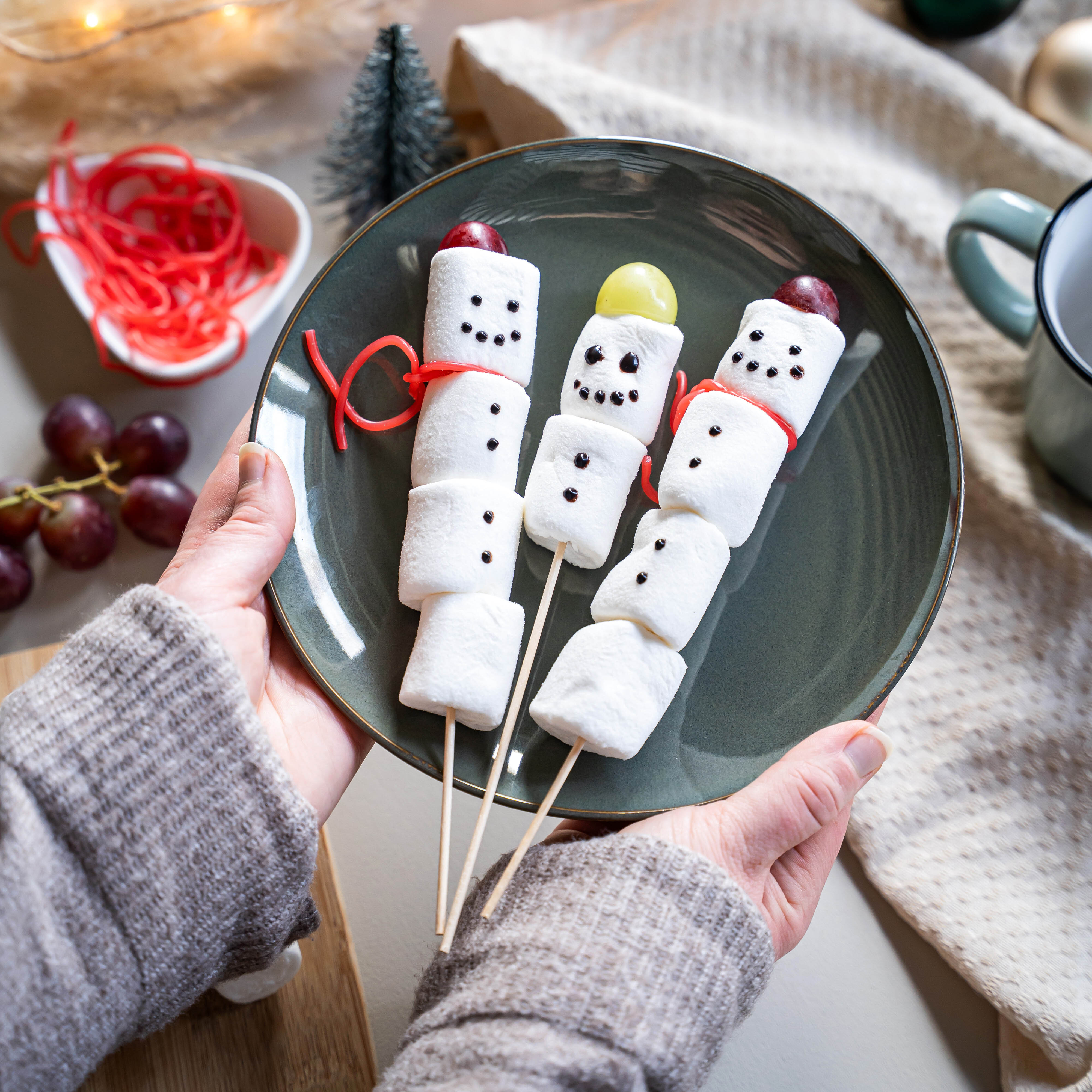 Drei Schneemänner aus weißen Marshmallows auf Holzspießen, mit Trauben als Köpfen und roten Bändern als Schals auf einem grauen Teller.