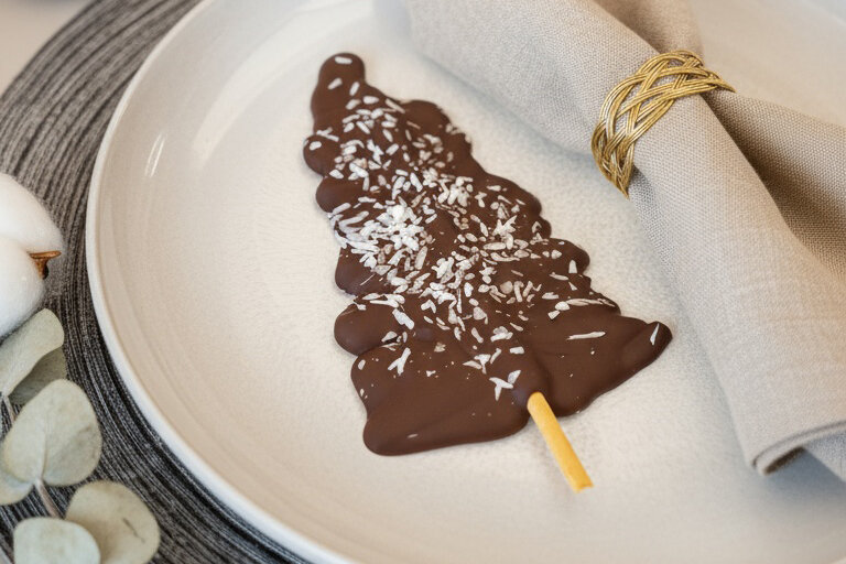 Schokoladen-Weihnachtsbaum mit Kokosraspeln auf weißem Teller, daneben graue Serviette mit goldfarbenem Serviettenring.