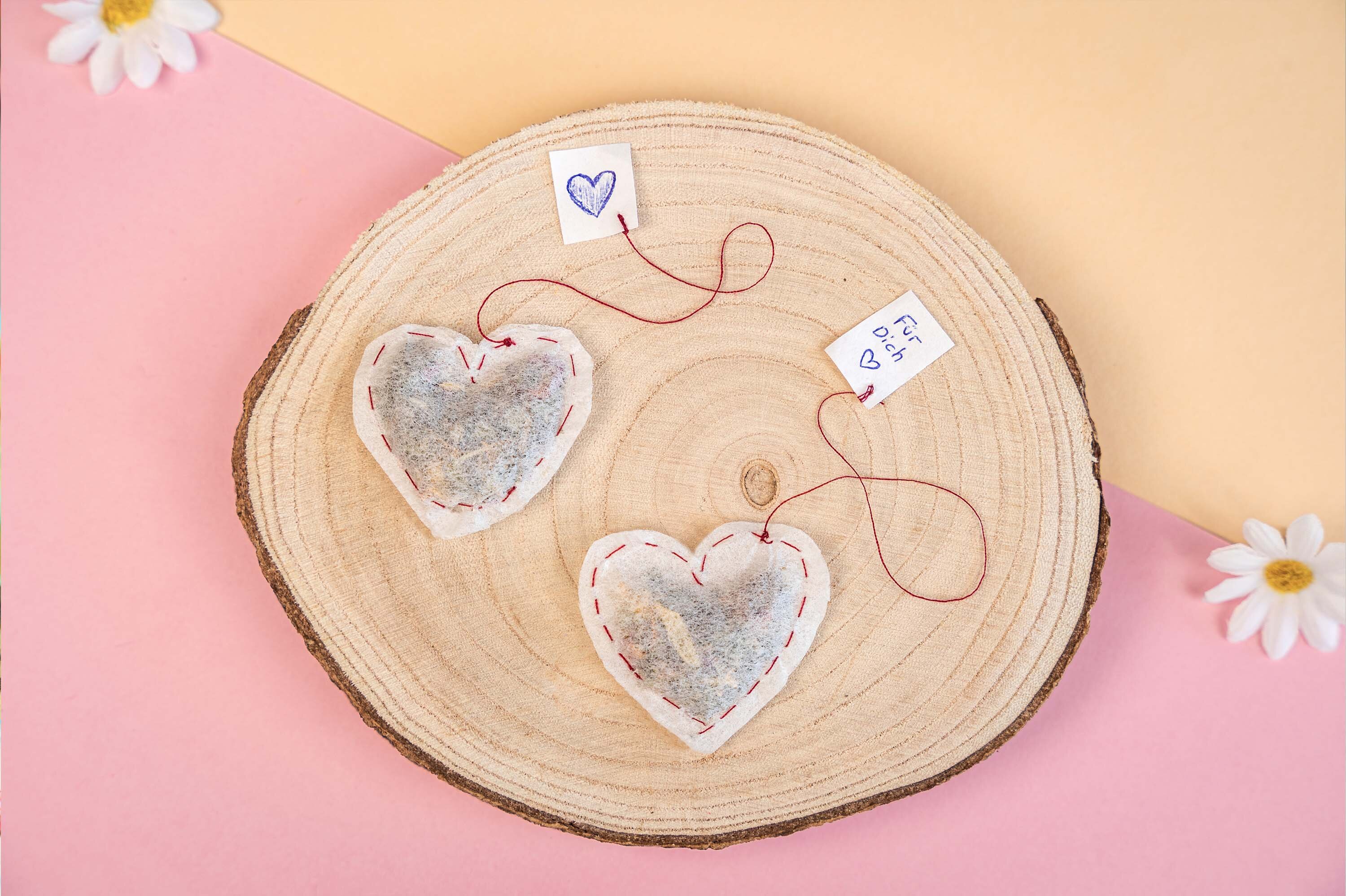 Zwei herzförmige Teebeutel mit roten Nähten auf einem Holzbrett. Jeder Beutel hat einen Faden mit einem kleinen Anhänger, einer mit einem blauen Herz und die andere mit 'P.S. ♥'