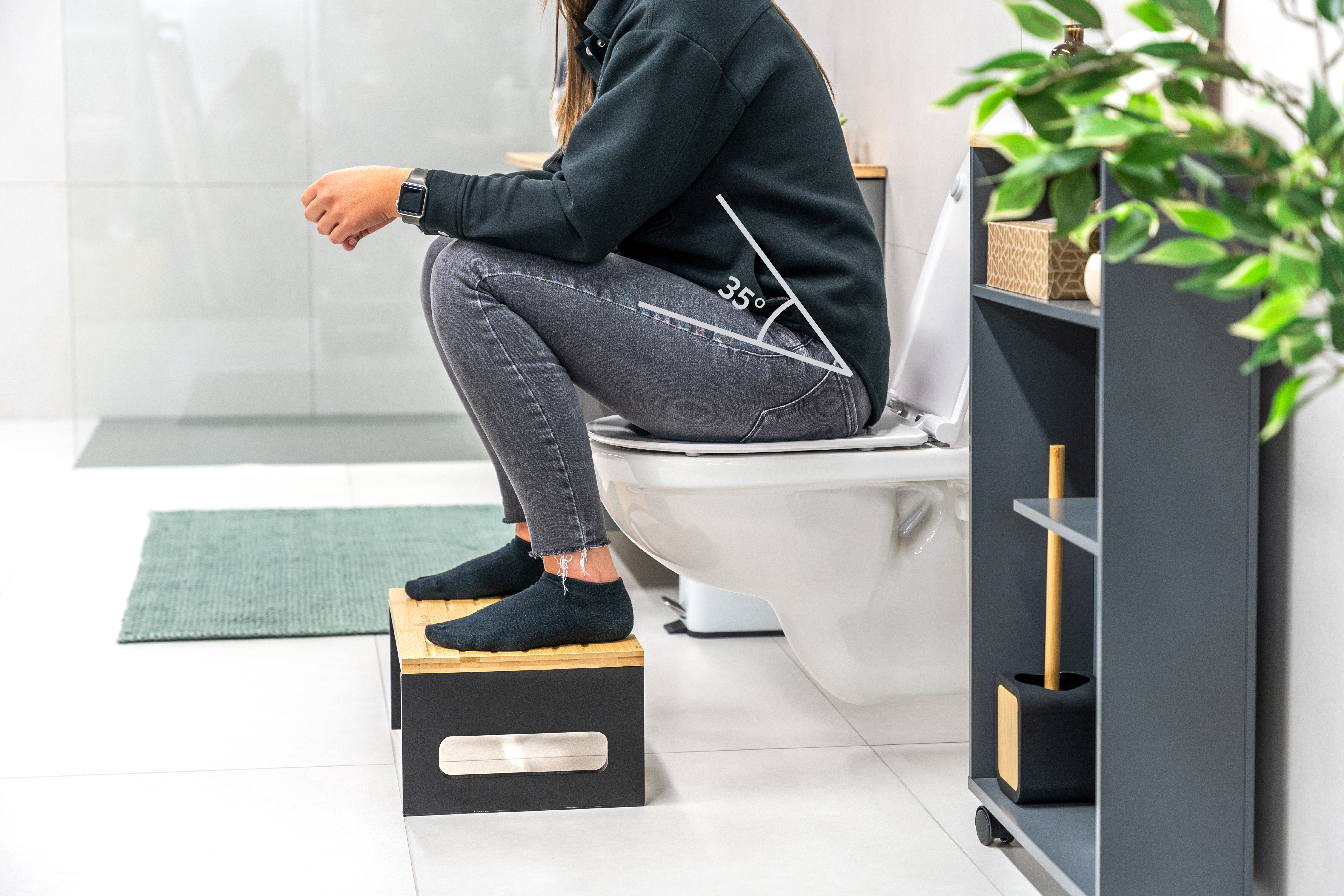 Eine Person sitzt auf einer Toilette und nutzt einen Hocker aus Holz, um die Füße darauf zu platzieren. Die Beine sind in einem Winkel von 35° angewinkelt. Im Hintergrund sind ein Handtuch und eine Pflanze sichtbar.