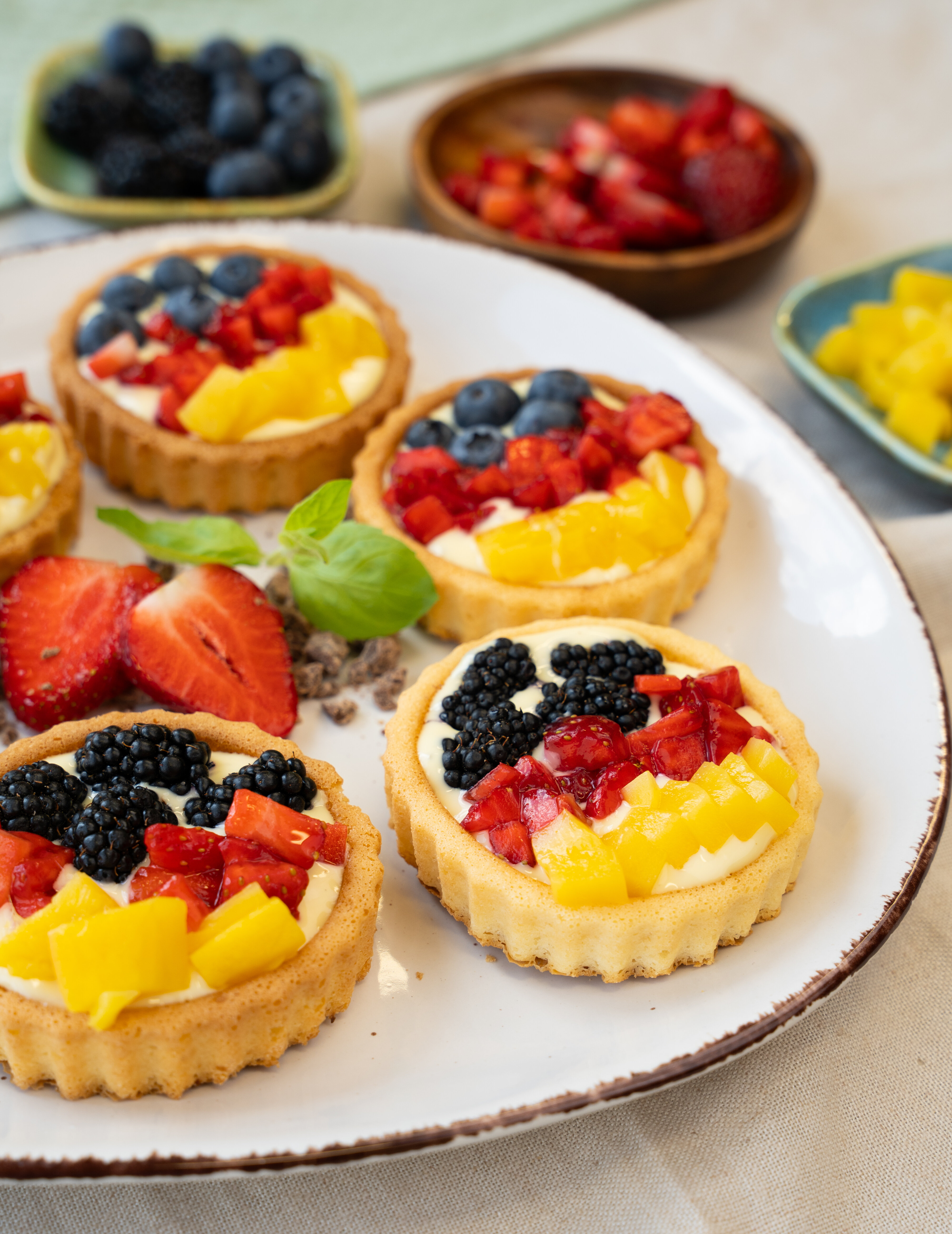 Mehrere kleine Törtchen mit Frischkäsecreme, belegt mit Brombeeren, Erdbeeren, Blaubeeren und Mangostücken auf weißem Teller