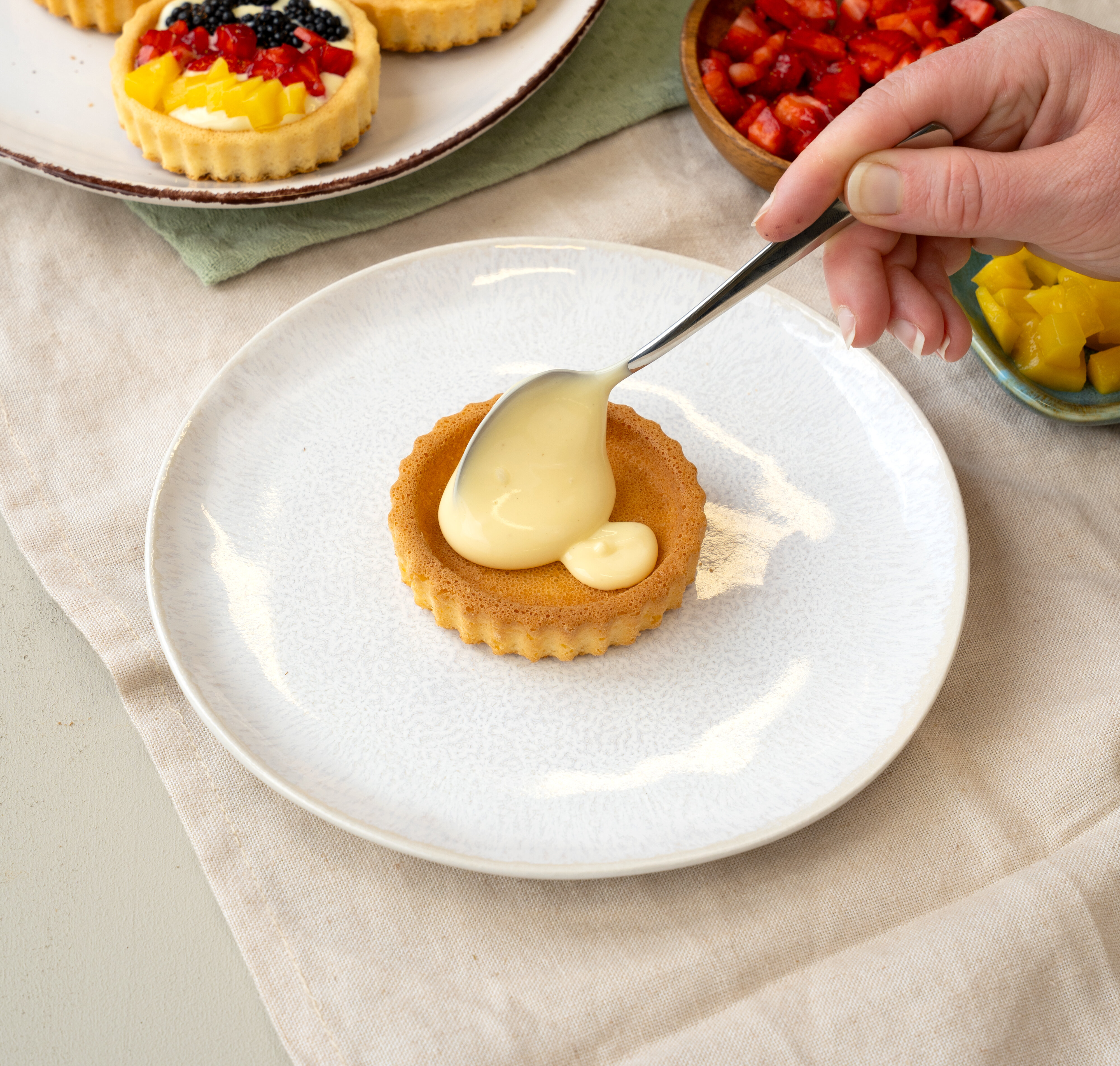 Hand, die mit einem Löffel eine helle Creme in eine kleine Tartelette-Form auf einem weißen Teller streicht