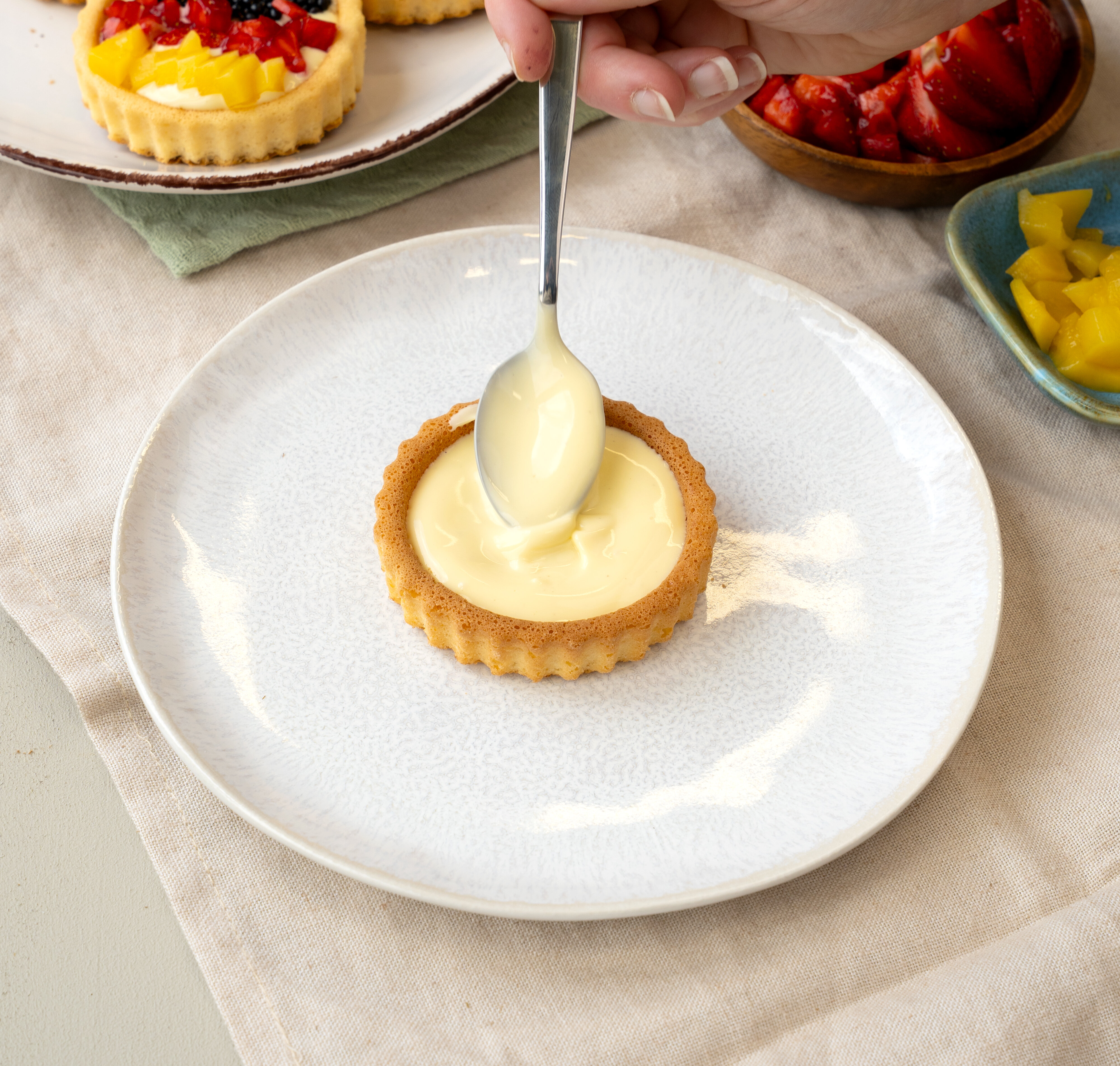 Hand, die mit einem Löffel eine helle Creme in eine kleine Tartelette-Form auf einem weißen Teller streicht