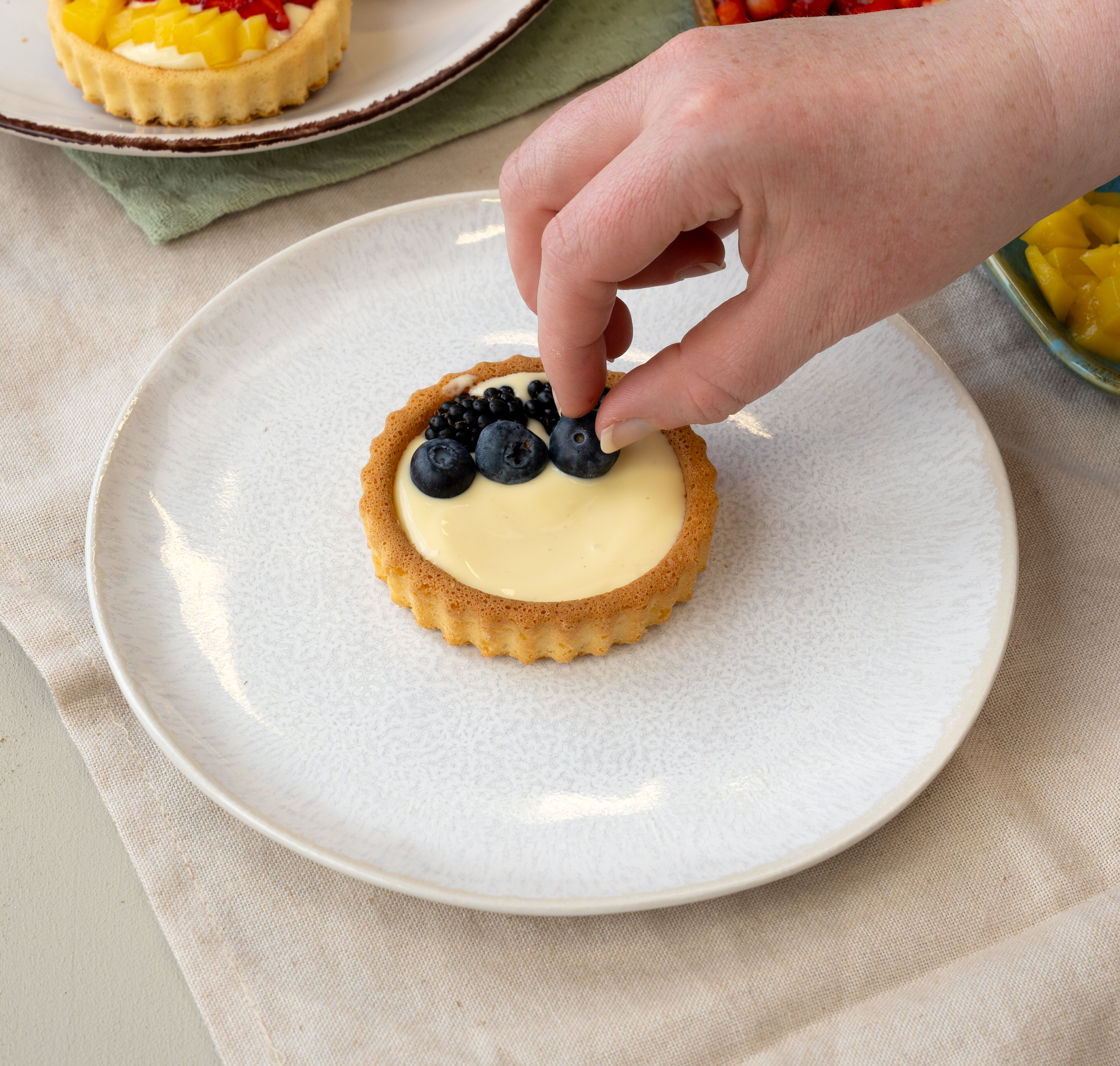 Hand legt Heidelbeeren auf eine kleine Tartelette mit Vanillecreme auf weißem Teller