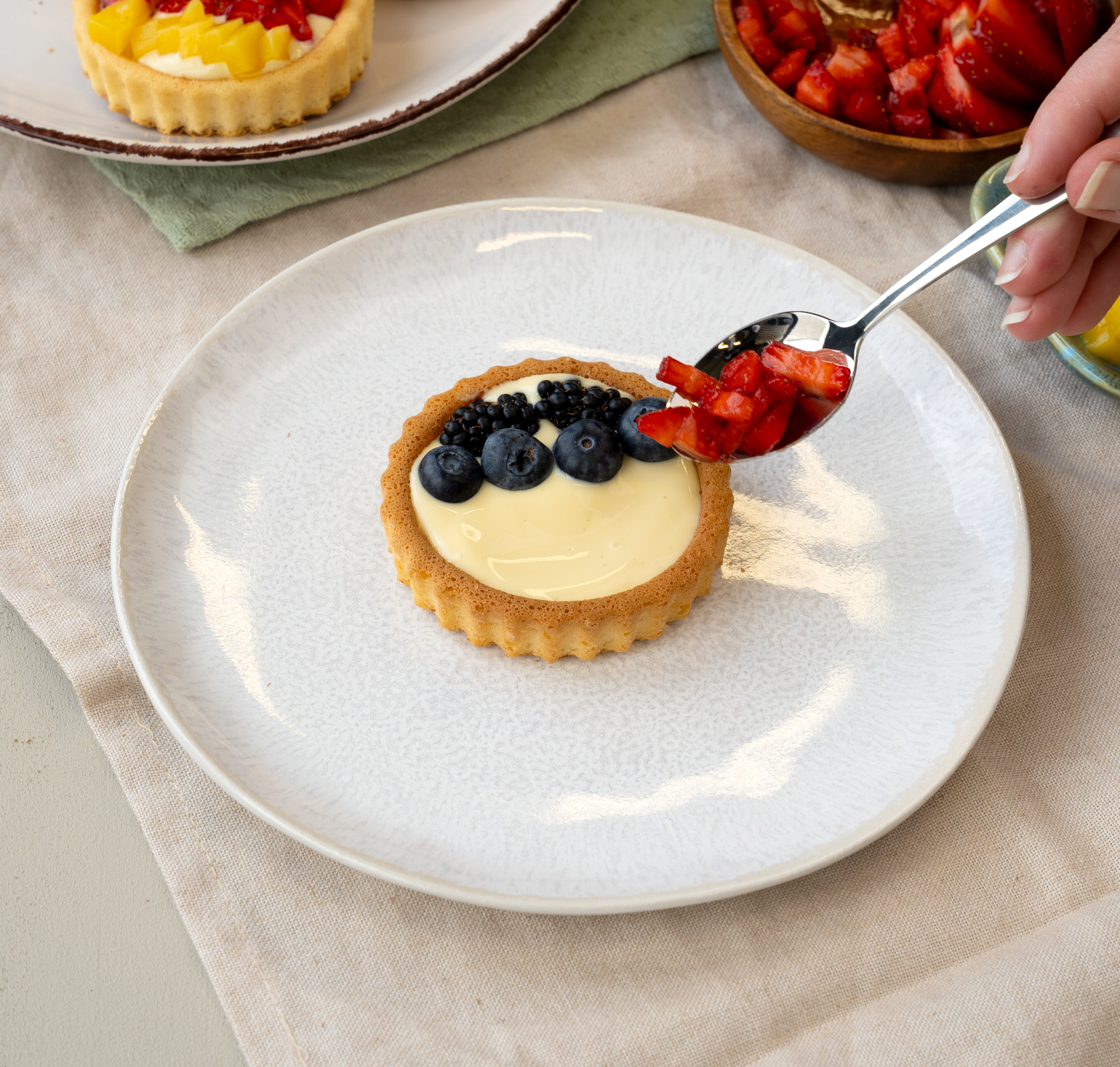 Tartelett mit Vanillecreme und Heidelbeeren auf weißem Teller, daneben wird mit einem Löffel geschnittene Erdbeere darauf gegeben