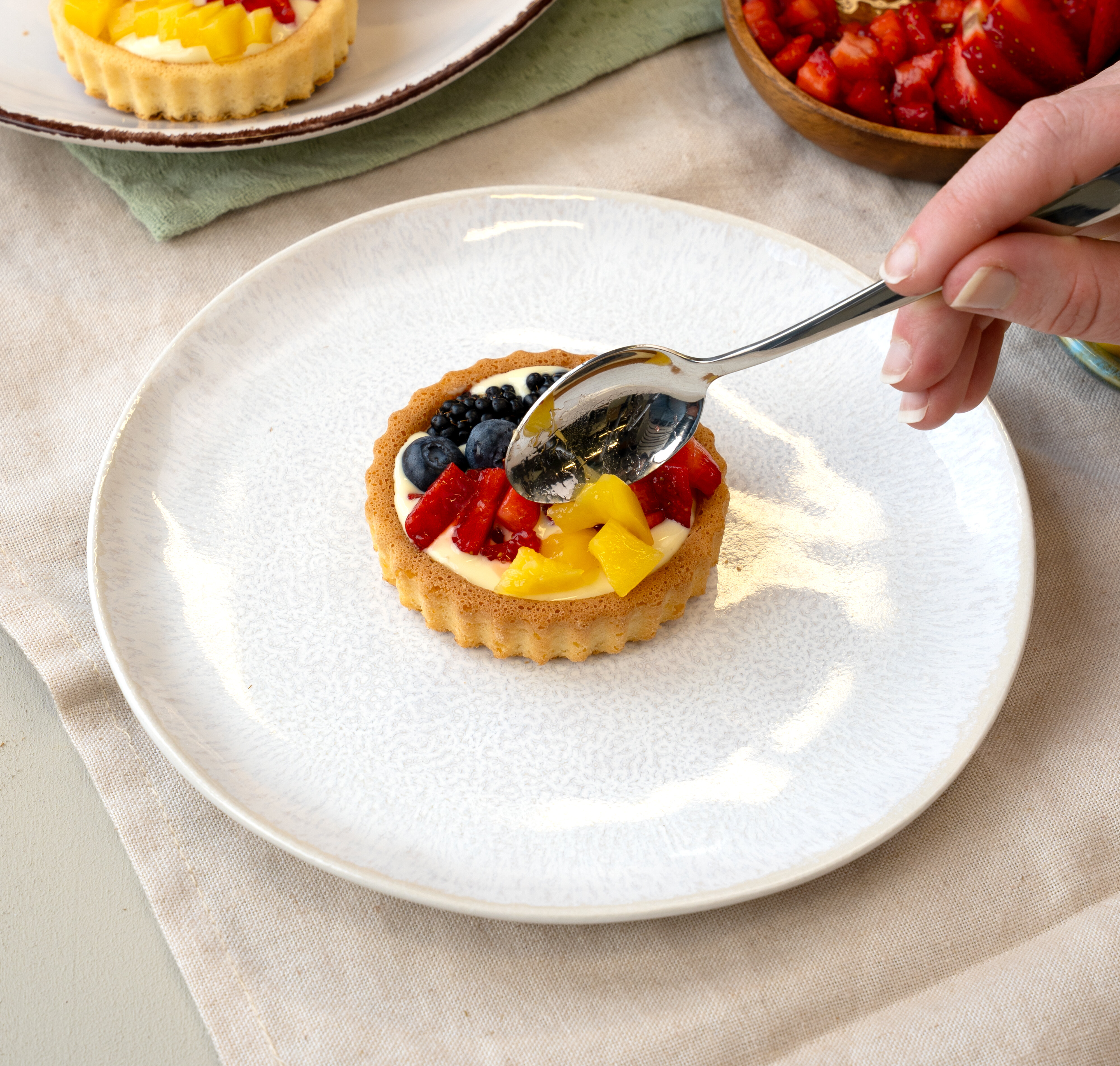 Hand mit Löffel dekoriert kleine Obsttartelette mit Blaubeeren, Erdbeeren und Mangostücken auf weißem Teller