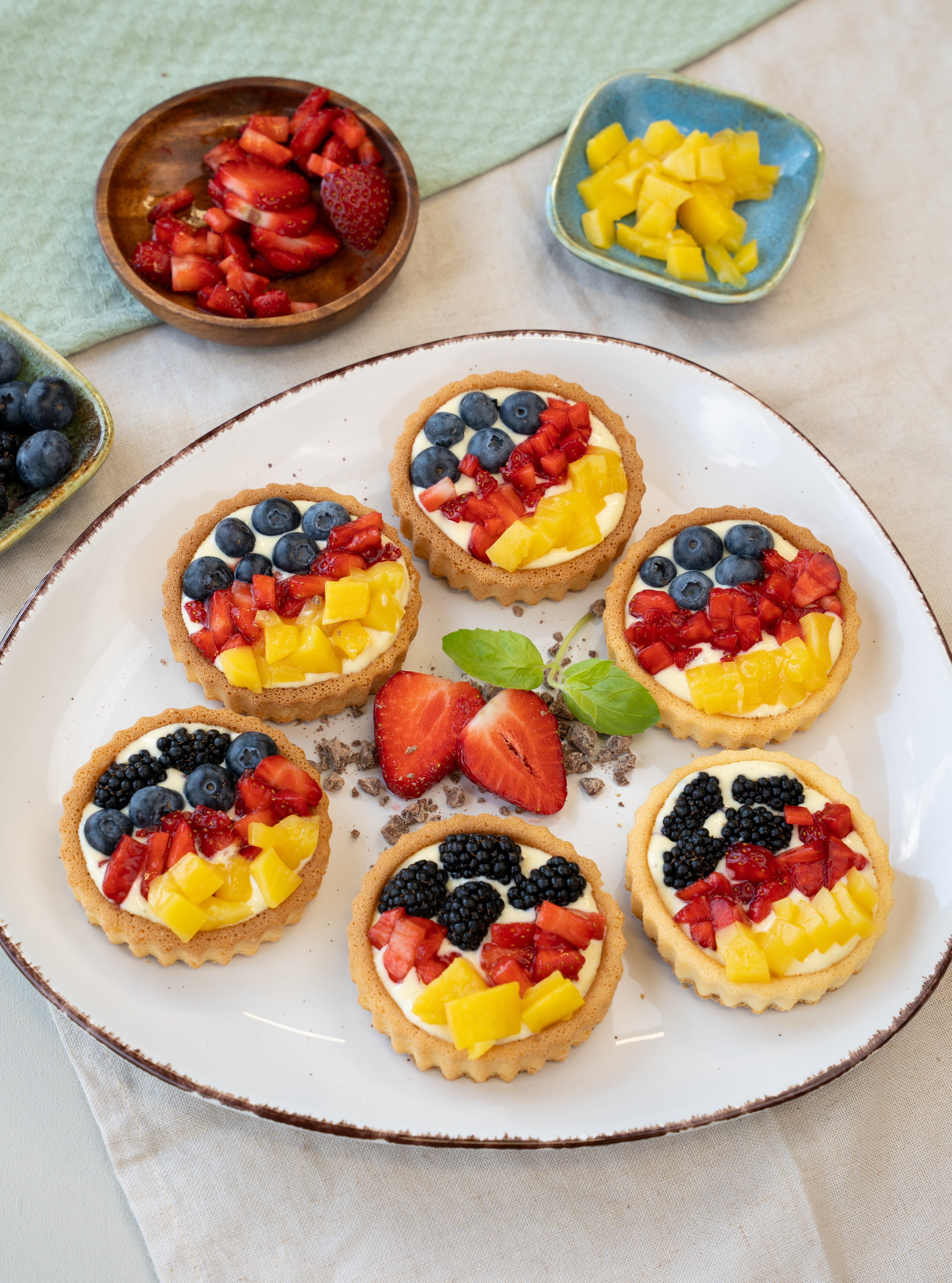 Sechs kleine Tartelettes mit Frischkäsecreme, belegt mit Blaubeeren, Brombeeren, Erdbeeren und Mangostücken auf einem weißen Teller, umgeben von kleinen Schalen mit zusätzlichen Früchten.