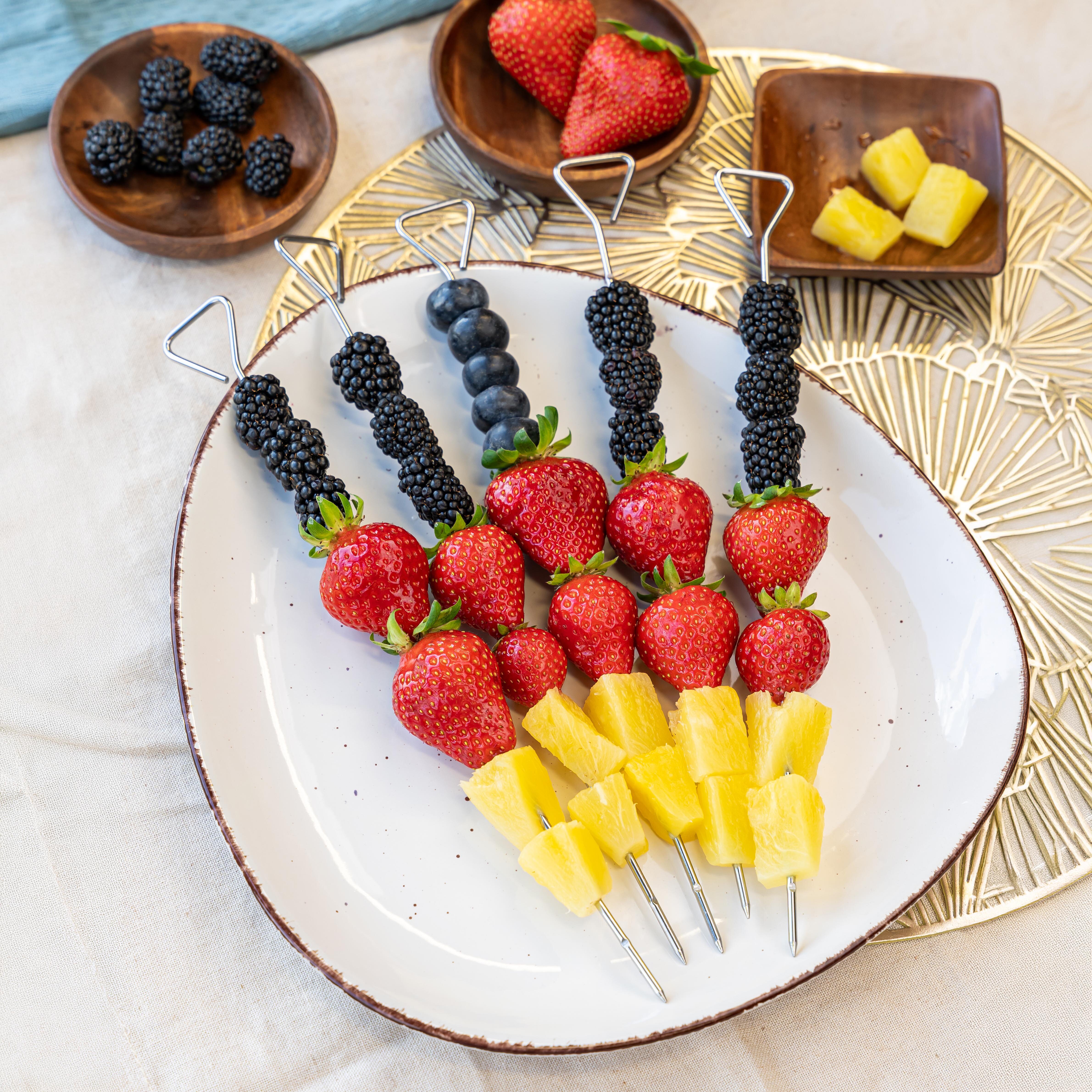 Auf einem ovalen, weißen Keramikteller liegen fünf Metallspieße, die jeweils mit einer Sorte Obst bestückt sind. Von links nach rechts sind die Spieße mit Brombeeren, Heidelbeeren, Brombeeren, Erdbeeren und Ananaswürfeln bestückt. Im Hintergrund stehen kleine Holzschälchen mit weiteren Brombeeren, Erdbeeren und Ananasstücken. Die Szene ist hell ausgeleuchtet und auf einem hellen Stofftuch arrangiert.