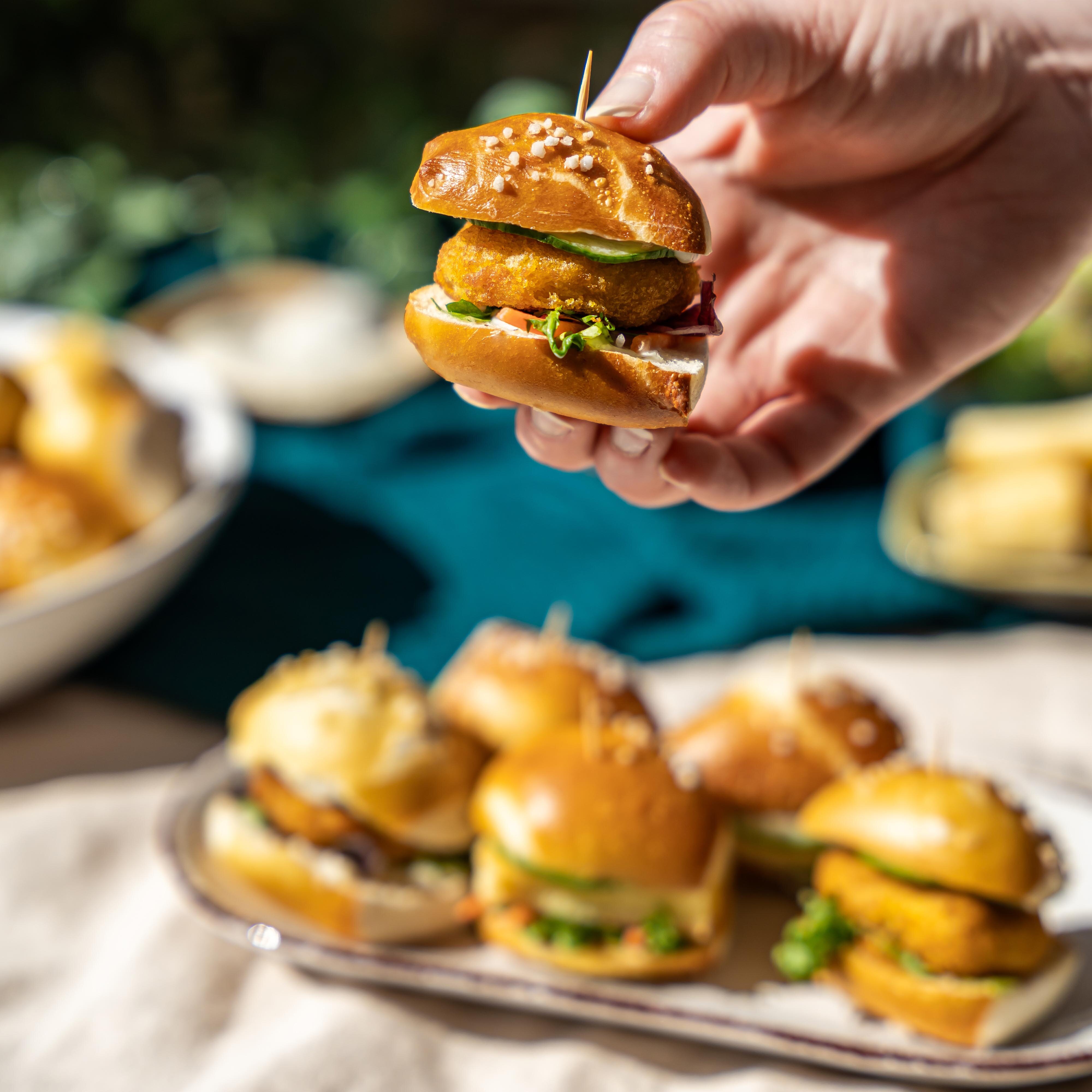 Eine Hand hält einen kleinen Burger mit Laugenbrötchen, Salat und einem panierten Patty. Im Hintergrund stehen weitere Mini-Burger auf einer länglichen Platte, daneben eine Schale mit weiteren Speisen. Die Szene ist hell ausgeleuchtet, der Fokus liegt auf dem Burger in der Hand.
