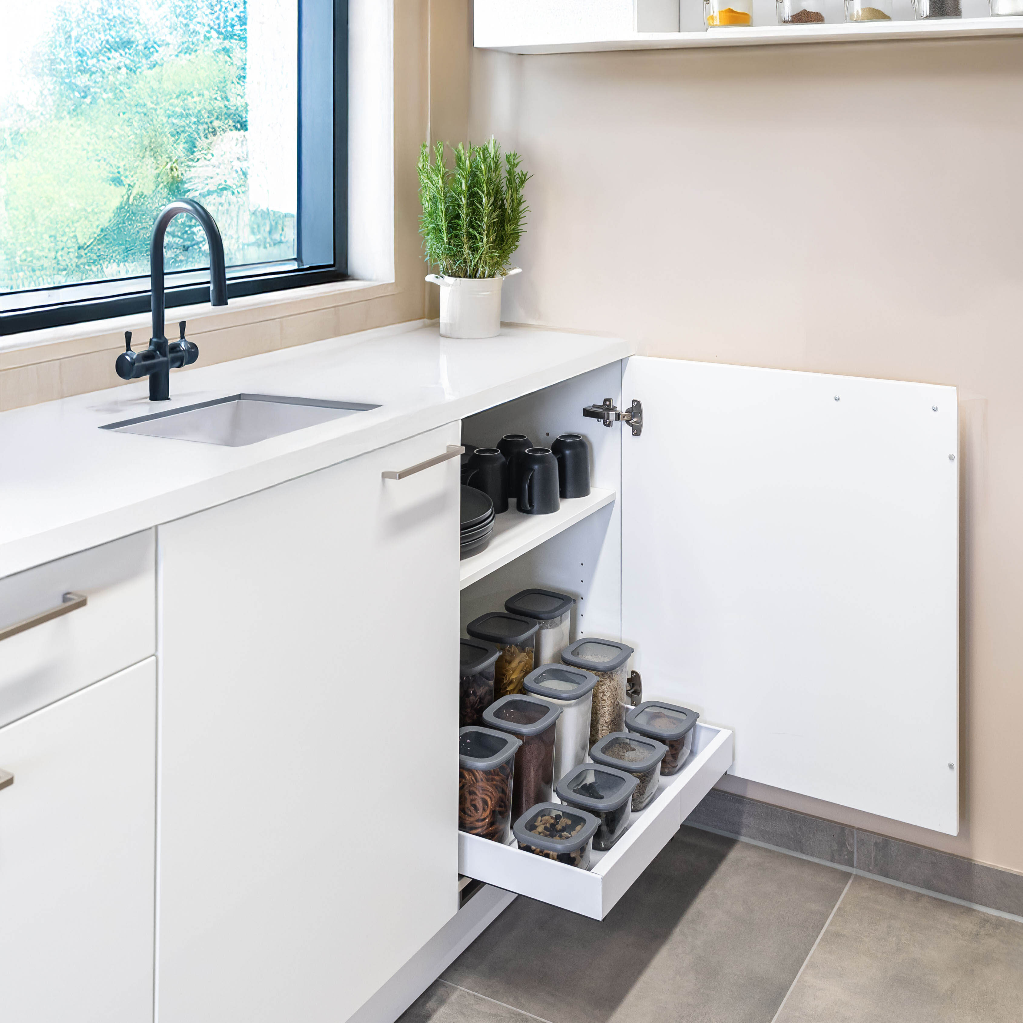 Weiße Küchenarbeitsplatte mit schwarzem Wasserhahn, Spüle, Kräutertopf und offener Schublade mit Vorratsbehältern und Geschirr.